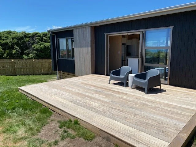 A wooden deck with two gray chairs outside a modern house with large windows and a sliding door under a blue sky.