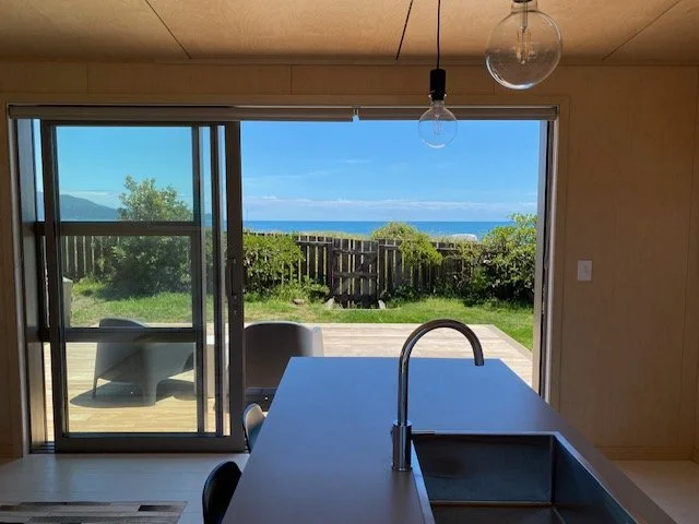 Interior view of a room with a kitchen island, including a modern sink and faucet, overlooking a backyard with a deck, greenery, and a view of the ocean and sky through sliding glass doors.