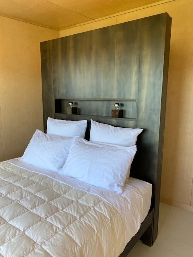 A bed with white pillows and a white quilt, with a tall, dark green headboard and built-in reading lights in a bedroom with beige walls.