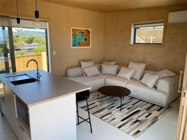 Living room with beige sofa, throw pillows, a small round black coffee table, patterned rug, and a kitchen island with a sink and stool. There is a sliding glass door and two windows, with wooden walls and ceiling.