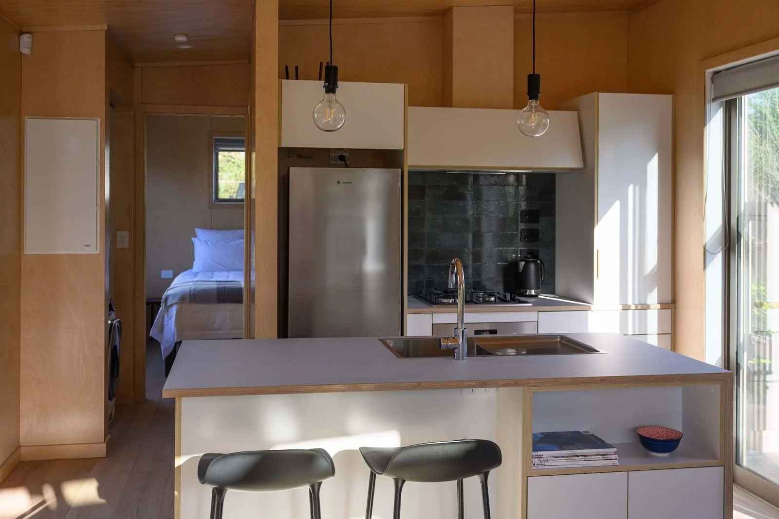 Modern kitchen with wooden cabinets, a white island with a sink, bar stools, and a view into a bedroom with a bed.