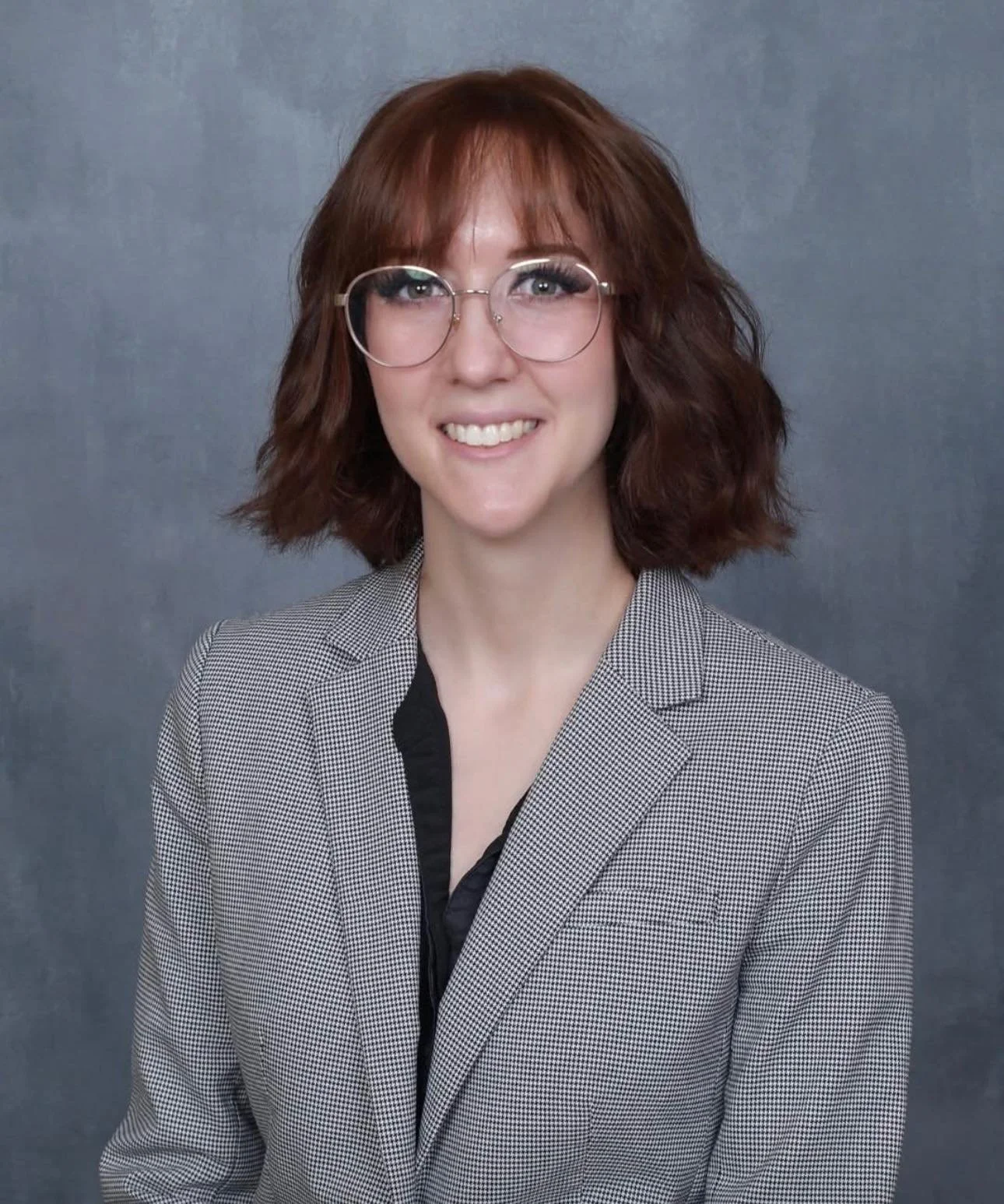 A woman with shoulder-length reddish-brown hair and glasses smiling at the camera, wearing a checkered blazer over a black top, standing against a gray background.