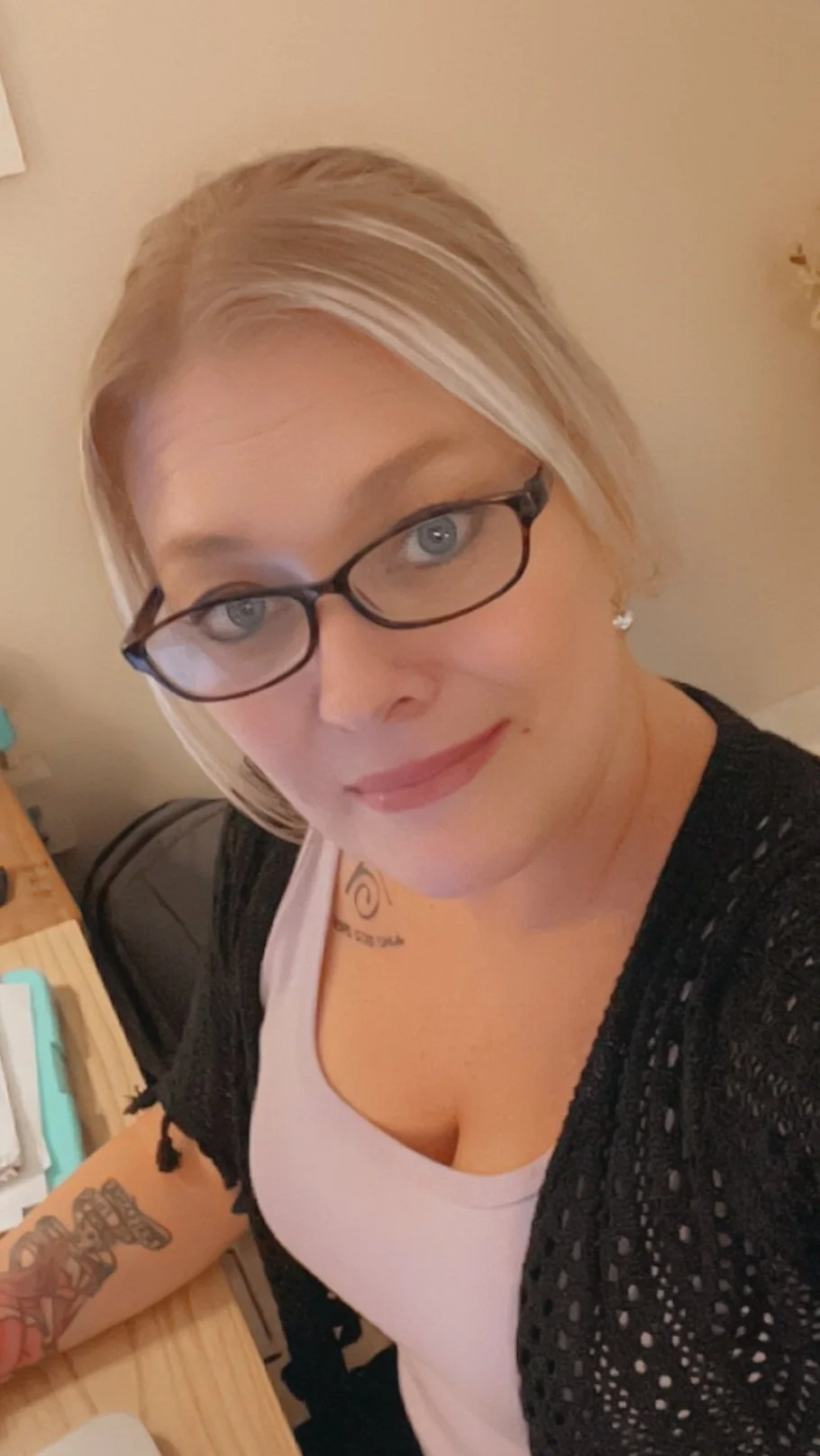 A woman with blonde hair, glasses, and tattoos on her arm, smiling at the camera, sitting at a wooden desk in a room with beige walls.