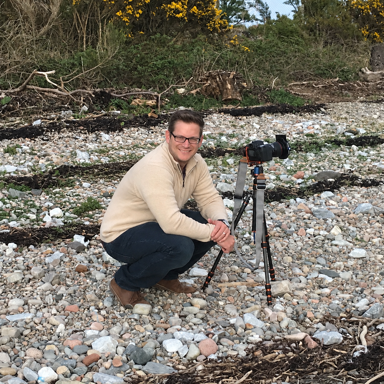 A man with glasses kneeling on a rocky beach beside a camera on a tripod, smiling at the camera. There are trees and bushes in the background.