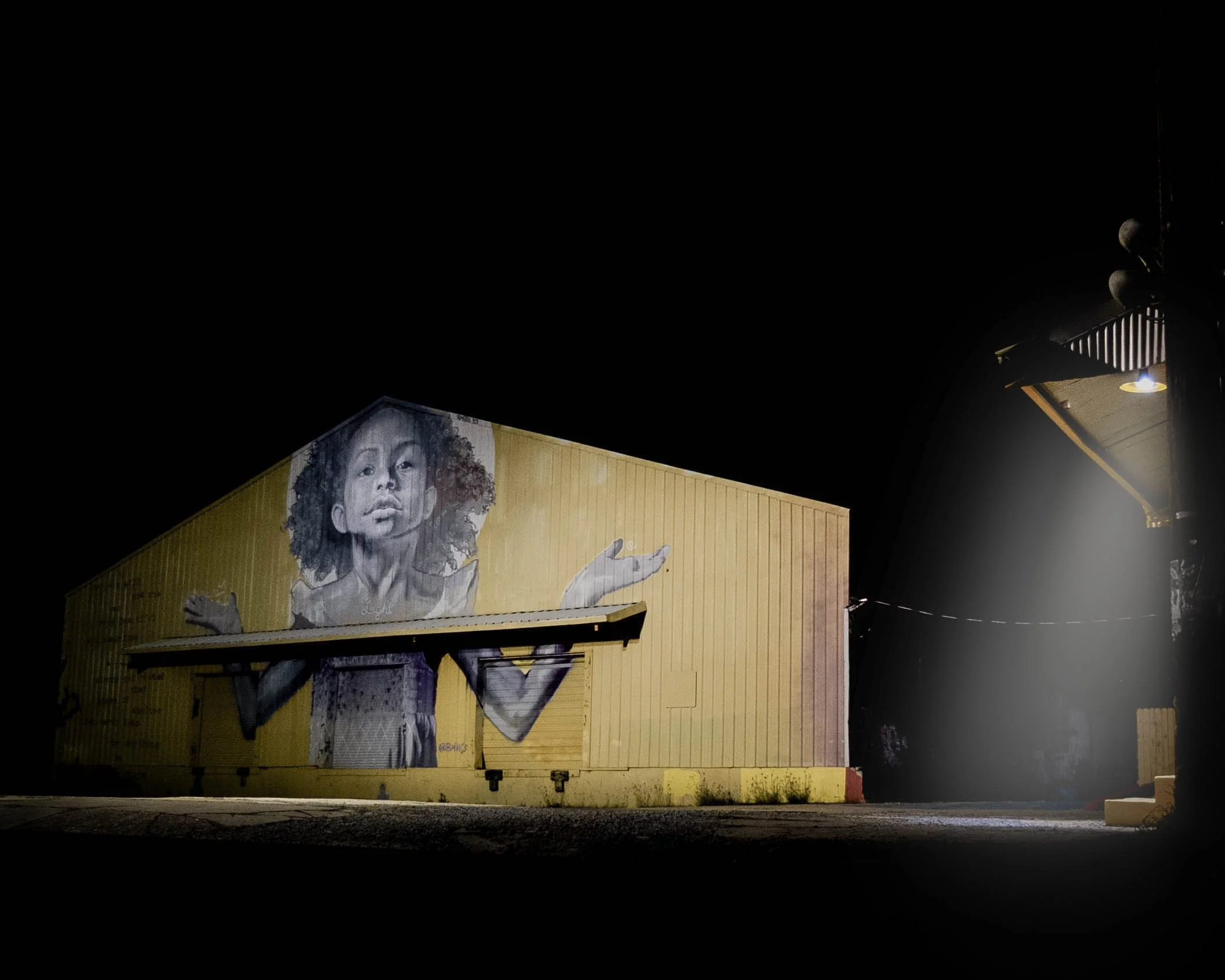 Nighttime mural of a child with curly hair and a serious expression on a yellow building wall.