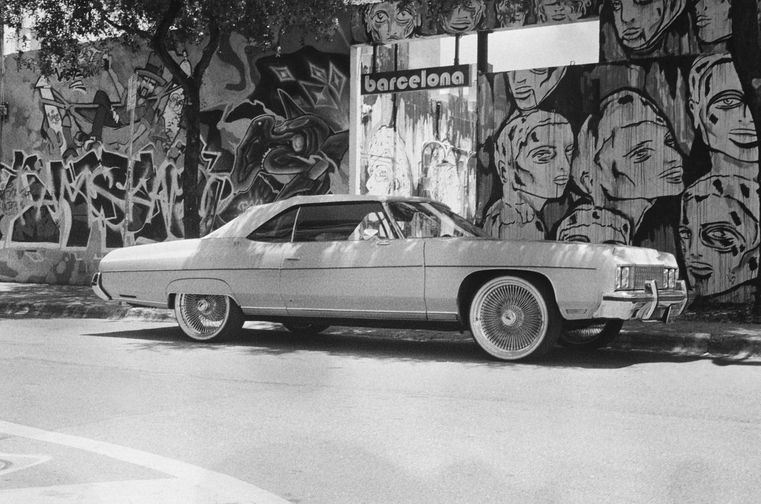 Black and white photograph of a classic car parked in front of a wall covered with colorful murals and graffiti, including faces and abstract designs, with a sign that reads 'Barcelona'.
