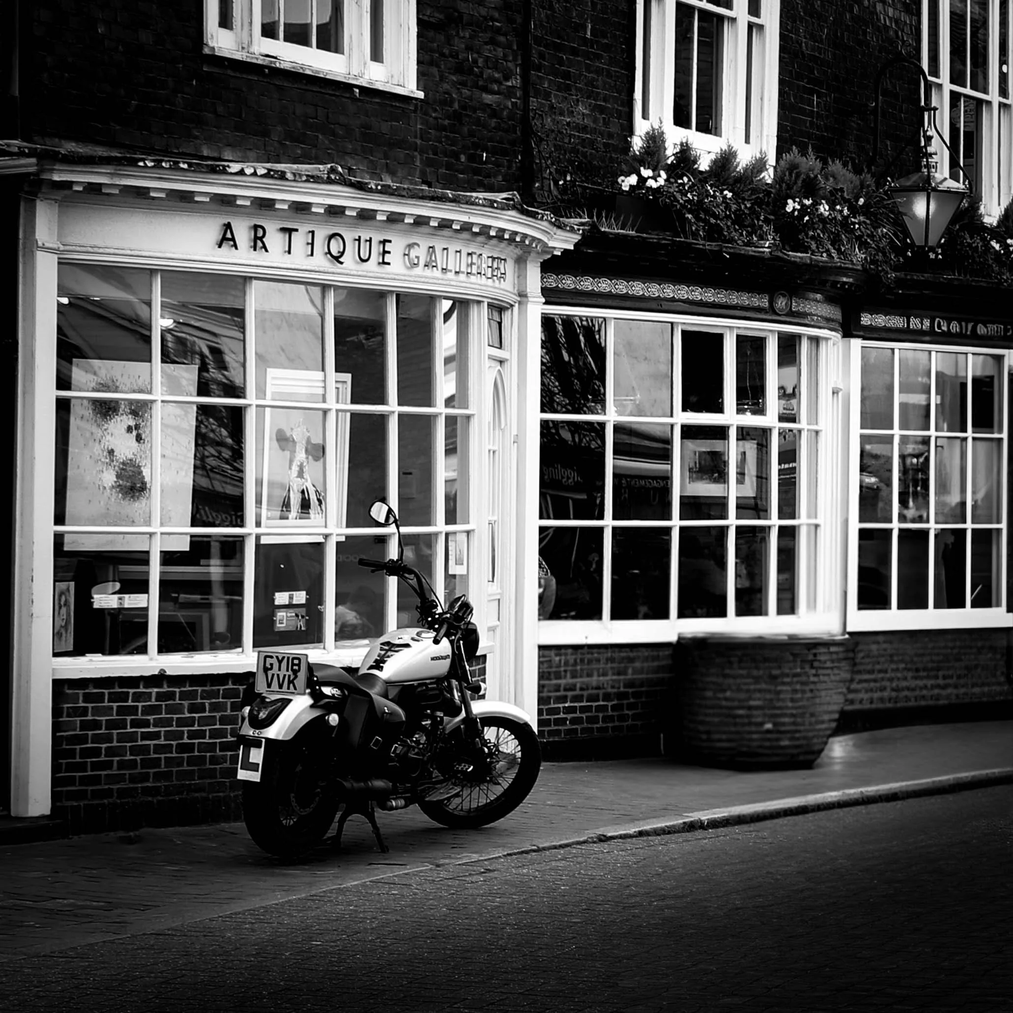 Black and white photo of a storefront window with the sign 'ARTIQUE GALLERY' and a motorcycle parked in front of it.