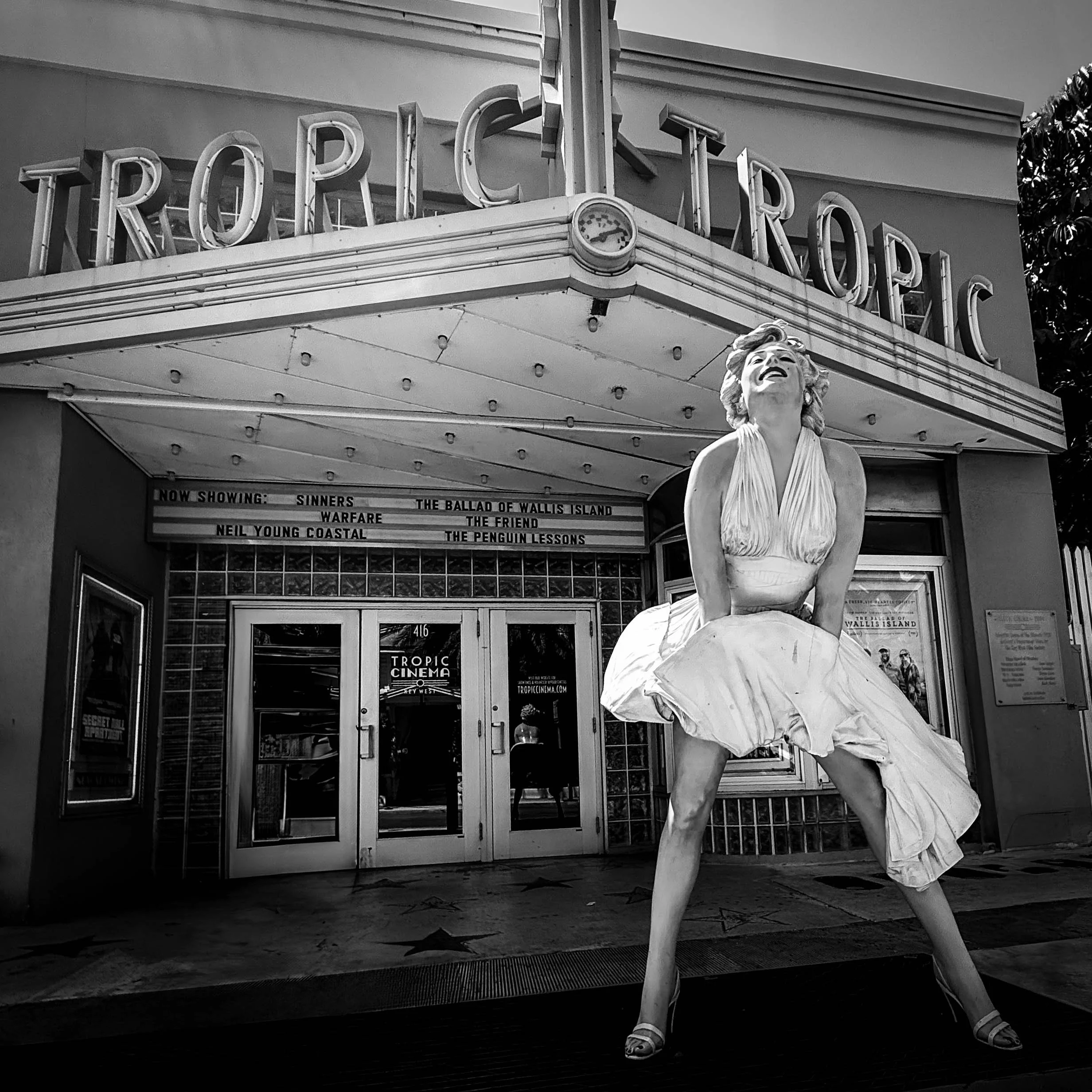 A black and white photo of a person in a dress with a short sleeve top walking past a theater marquee with white letters that spell 'TROPFIC'. The person is holding a puffy purse and wearing sandals. There is a classical statue of a woman in front of