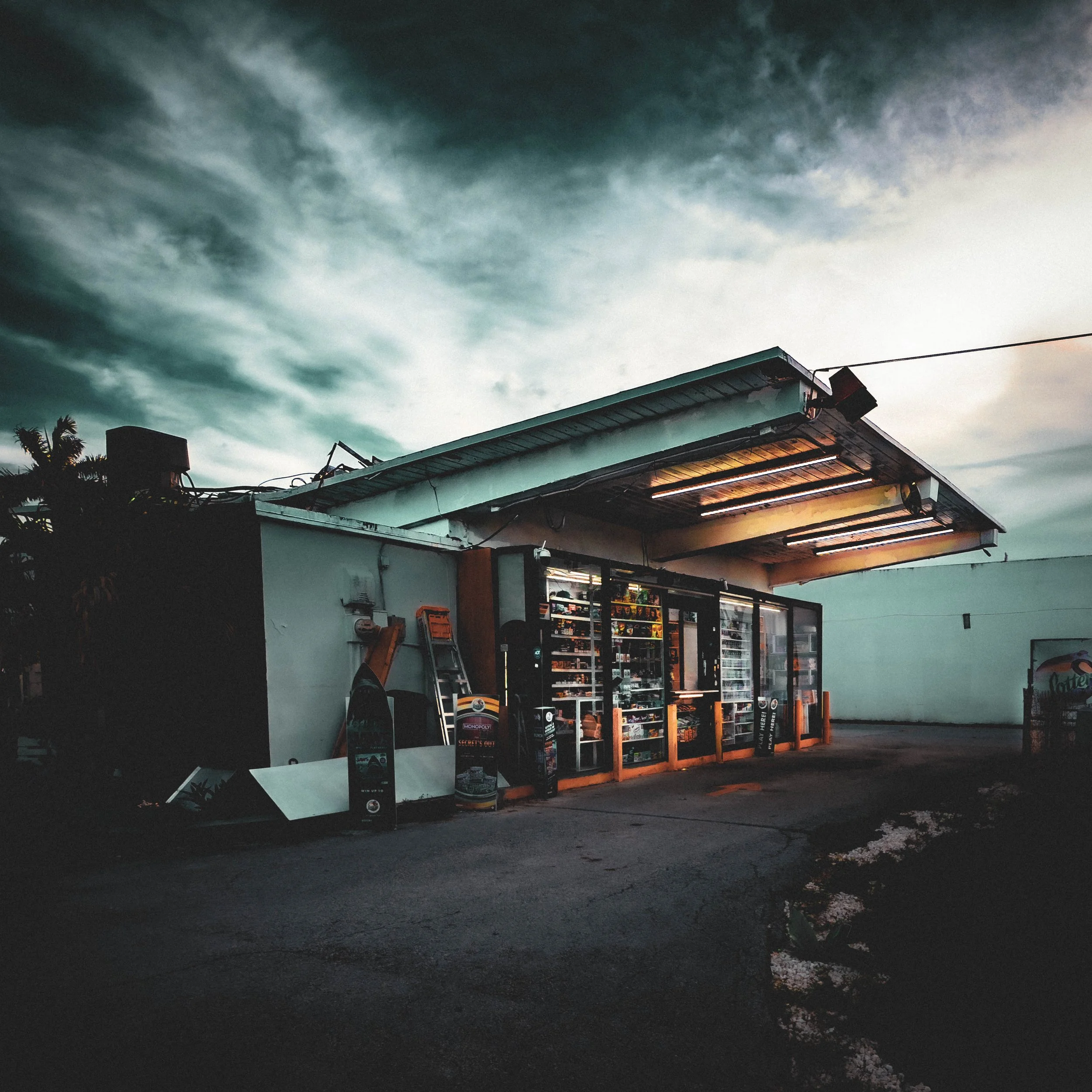 A convenience store with illuminated signage and shelves of products, located under a dramatic cloudy sky during dusk or early evening.