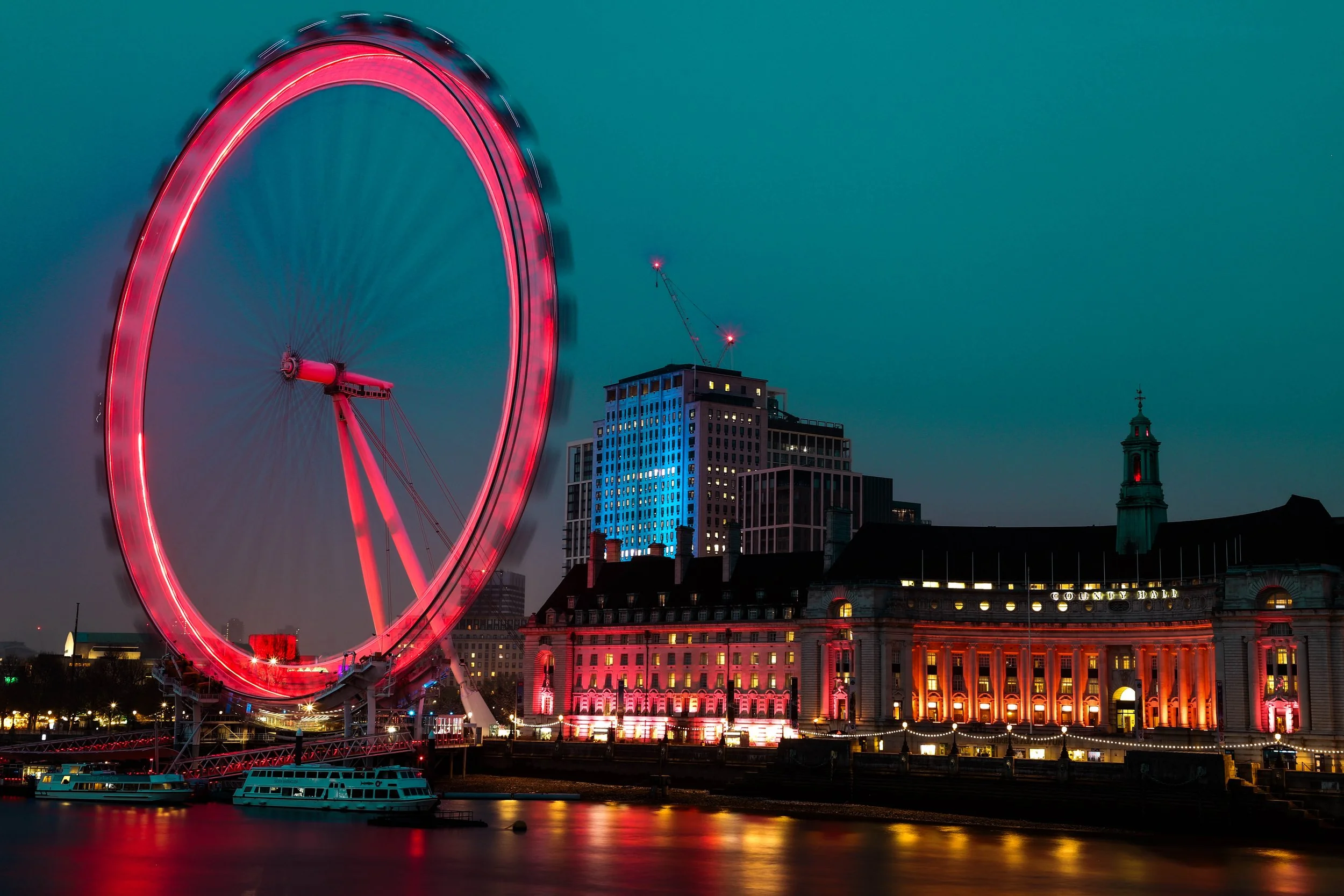 Nighttime cityscape with a brightly lit ferris wheel spinning, reflecting red and pink lights on the water below. Tall buildings with illuminated windows are in the background, and boats are docked along the waterfront.