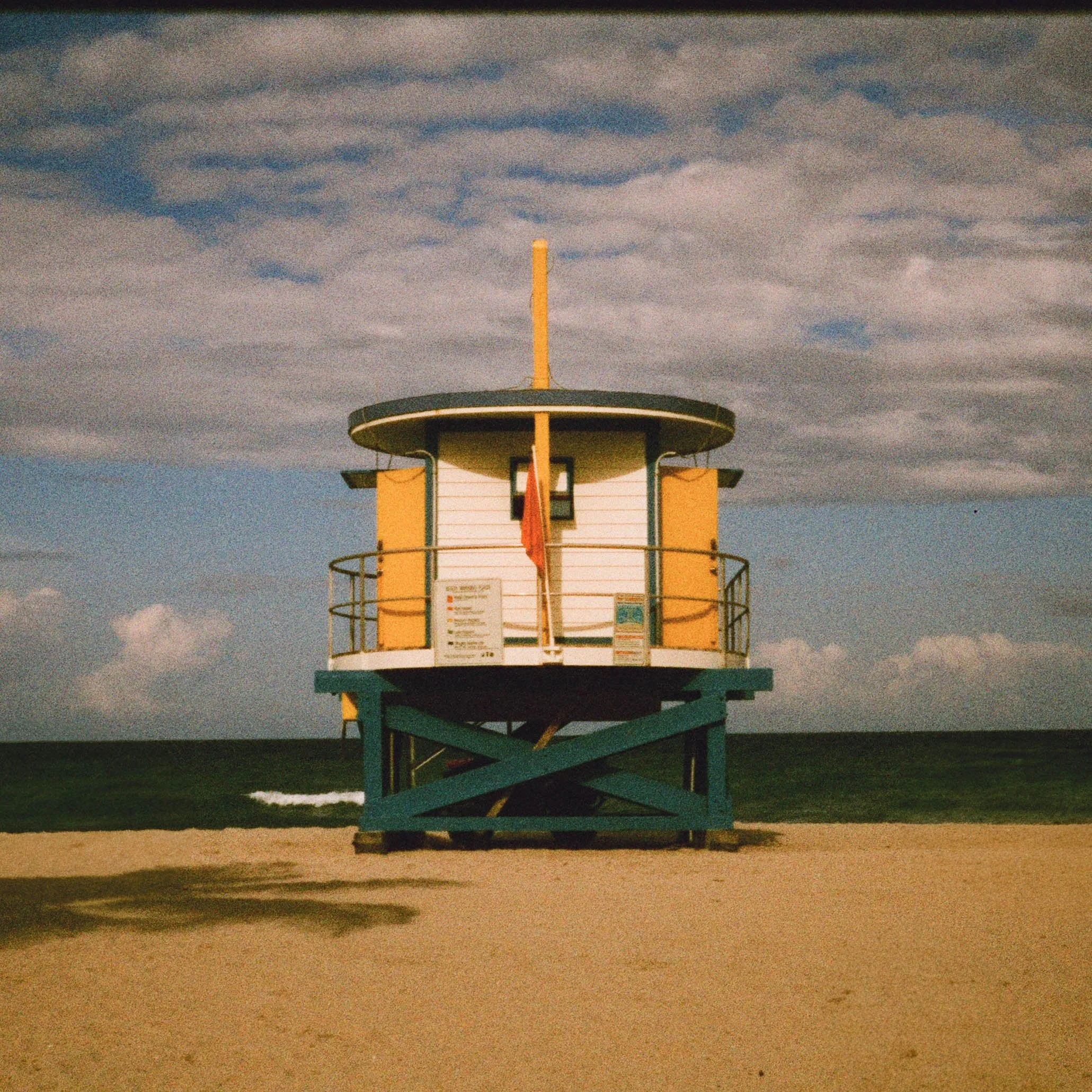 A vintage lifeguard tower painted in yellow and blue, standing on a sandy beach under a partly cloudy sky.