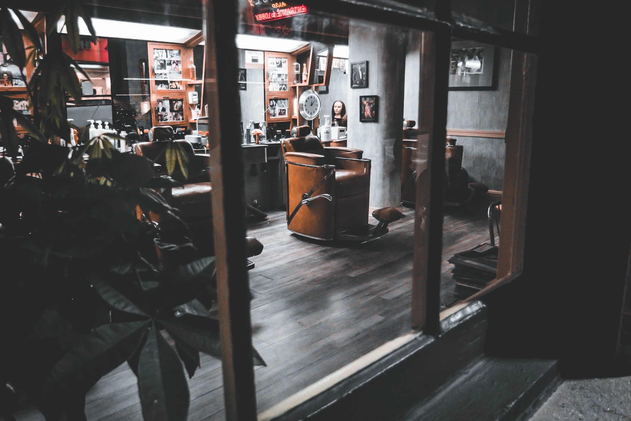 Interior of a barbershop with barber chairs, framed photos, a clock, and a mannequin head, viewed through a window.