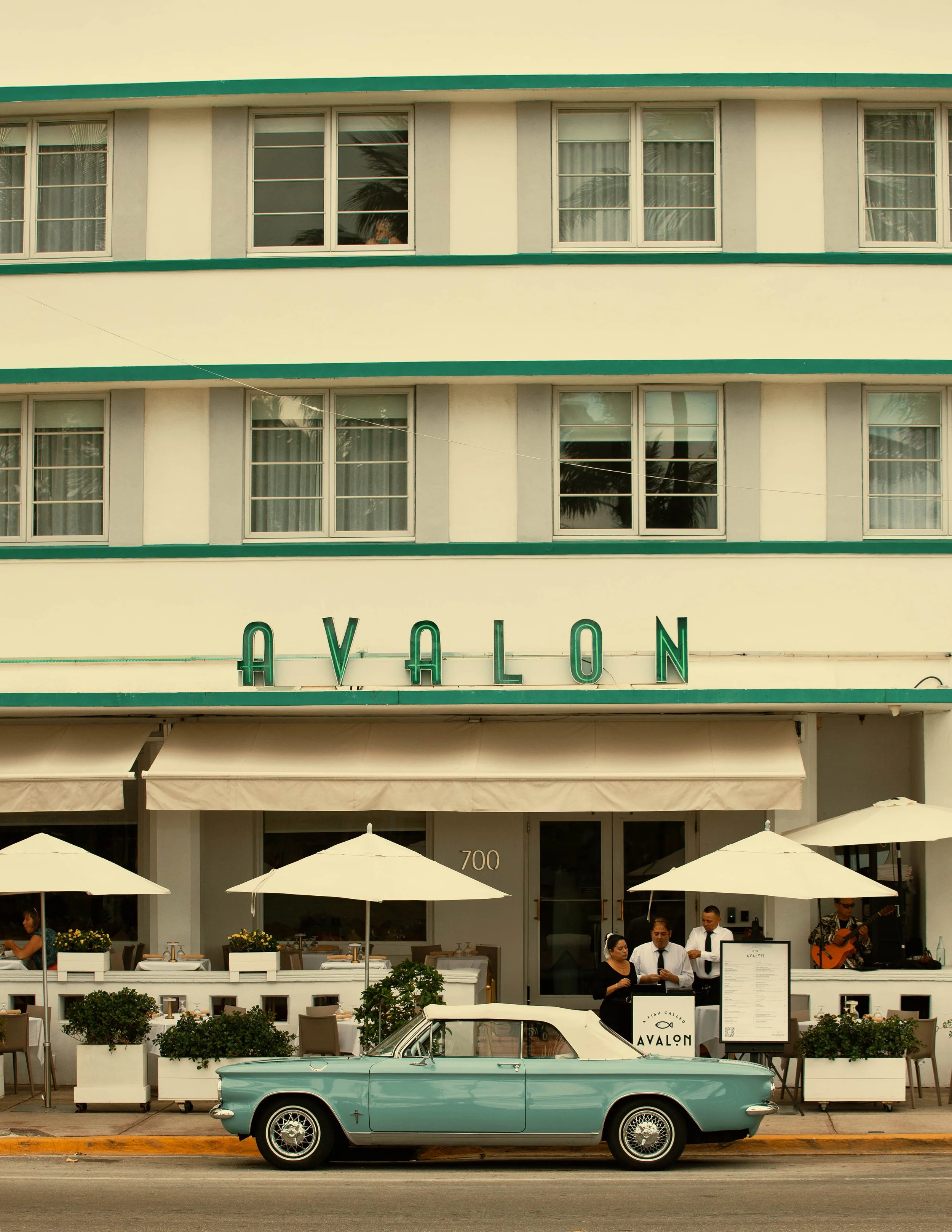 A classic blue convertible car parked on the street in front of a restaurant with outdoor seating, white umbrellas, and people dining, with a multi-story building in the background.