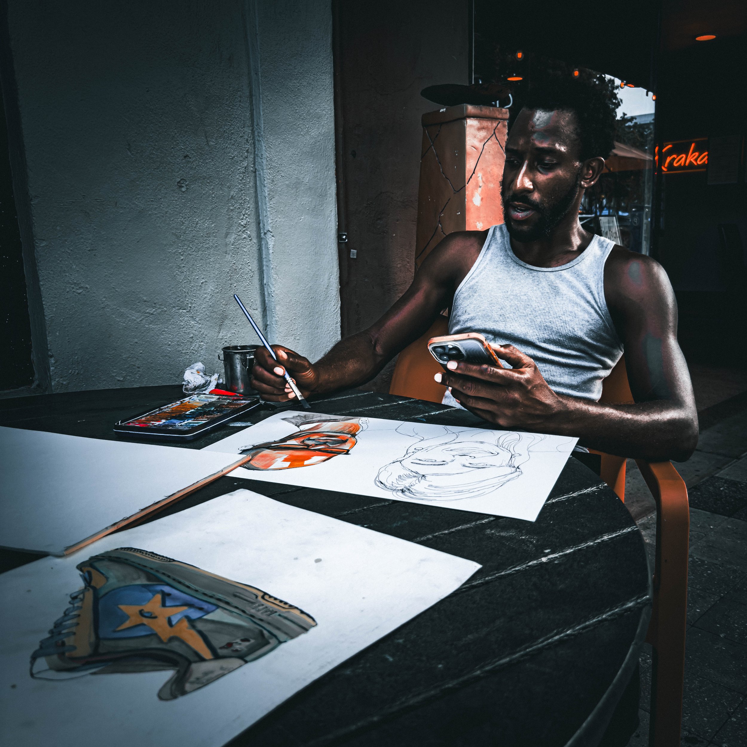 A man sitting at a table with art supplies, including colored markers, paper with sketches, and a sketchbook, in a dimly lit space with a textured wall and a neon sign in the background.