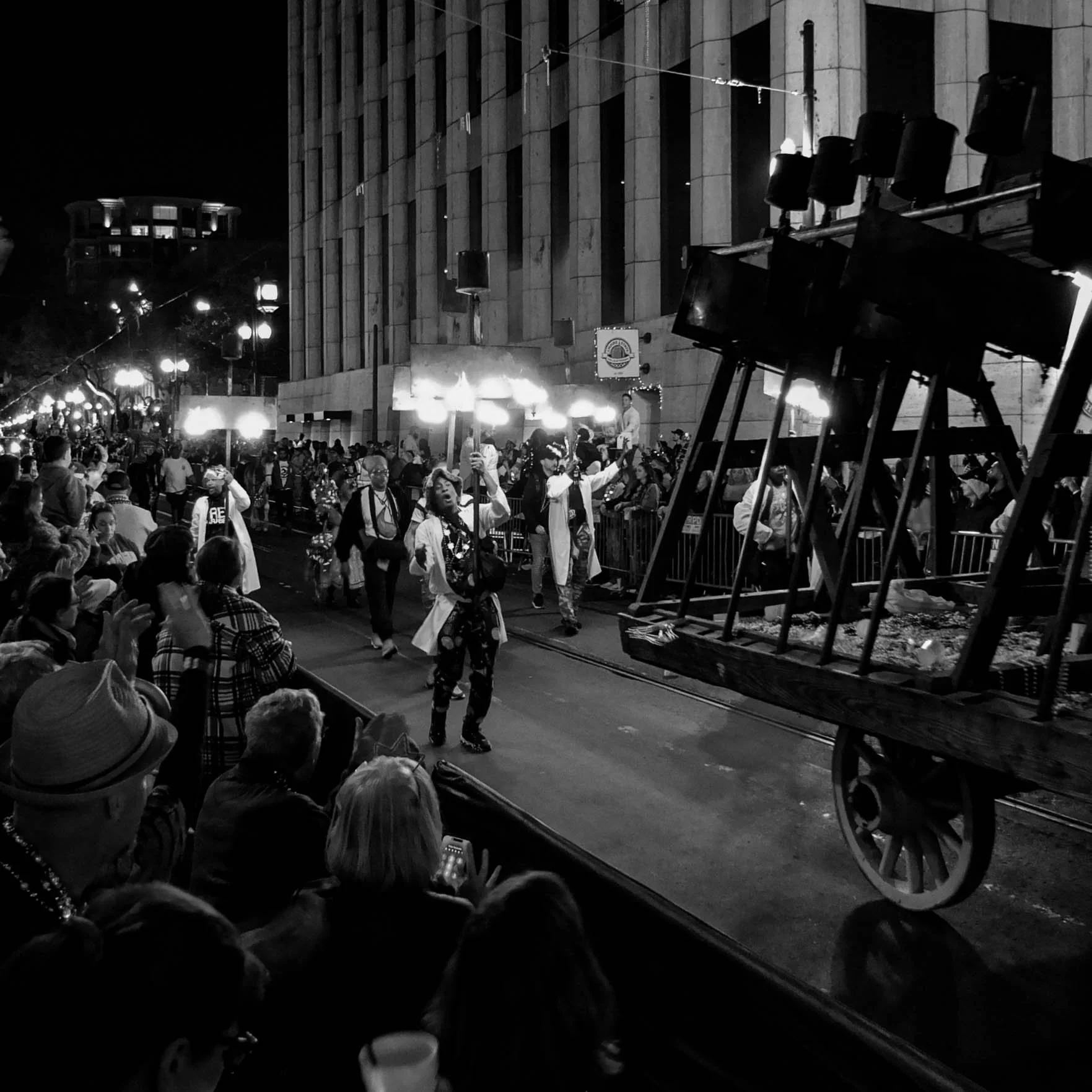 People gathered at night in front of a building with bright lights and a large screen, participating in a parade or celebration. Some individuals are holding signs, and there are staged speakers or equipment set up on the side.