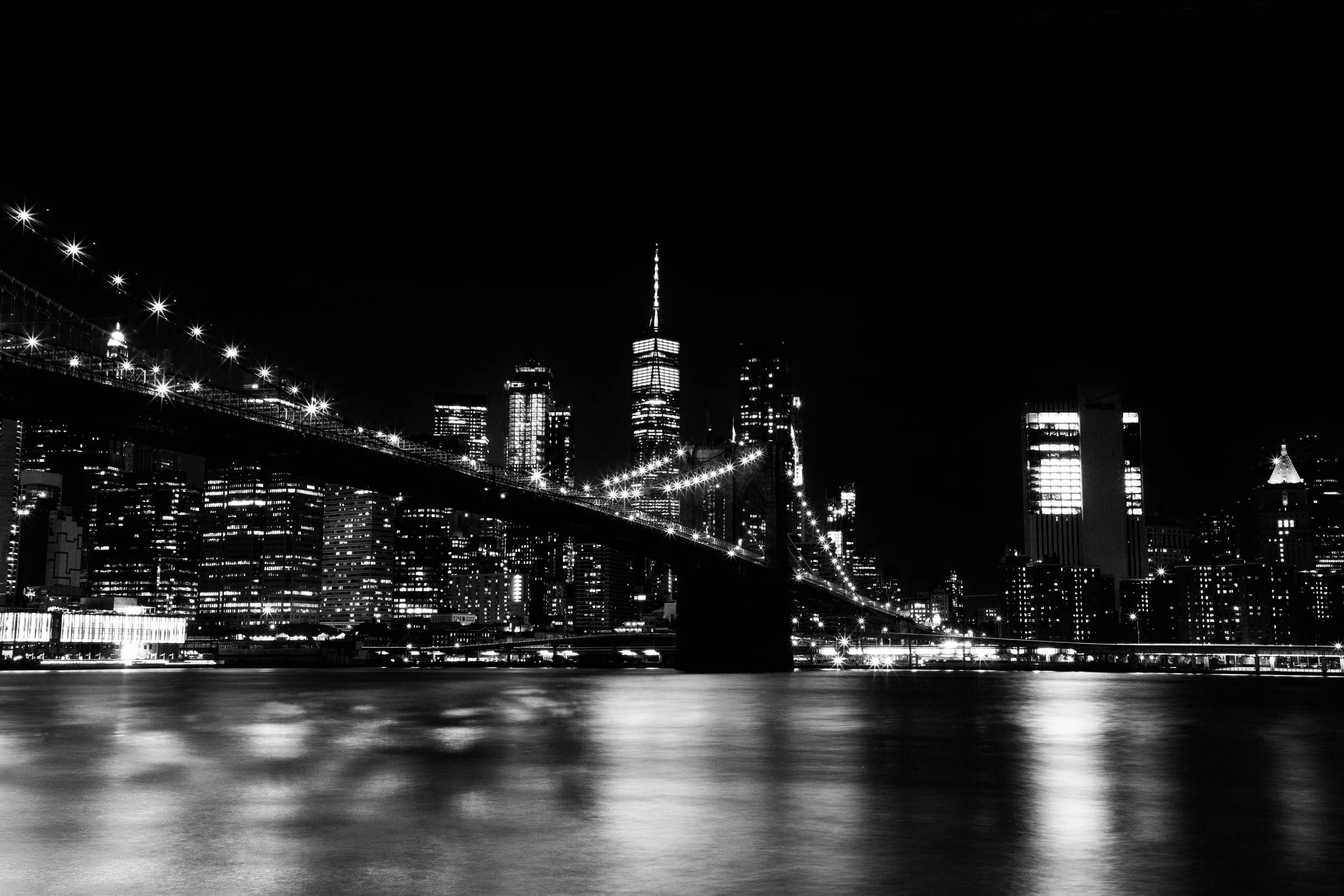 A black and white nighttime cityscape of New York City with illuminated buildings, including the One World Trade Center, viewed from across the water with reflections on the surface.