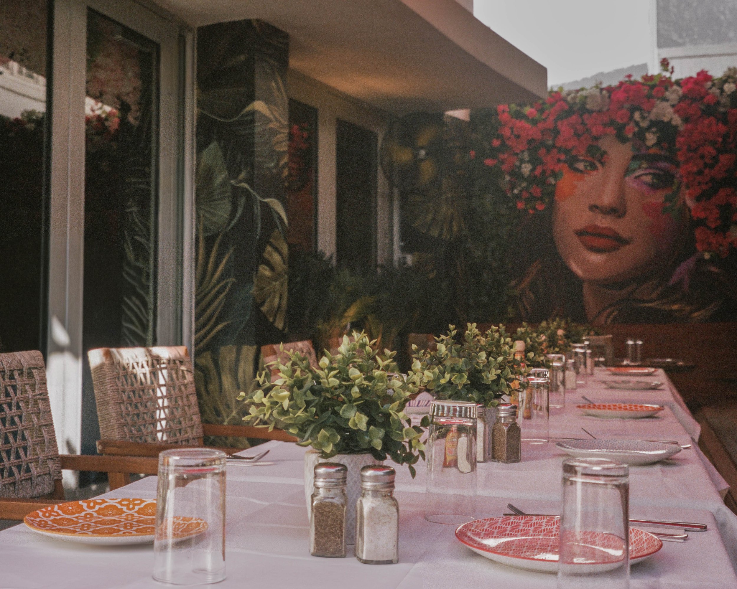 Outdoor dining area with a long table set with plates, glasses, and small jars of spices. The table is decorated with a green leafy plant centerpiece. There is a large mural of a woman's face with vibrant flowers as her hair on the wall behind, and g