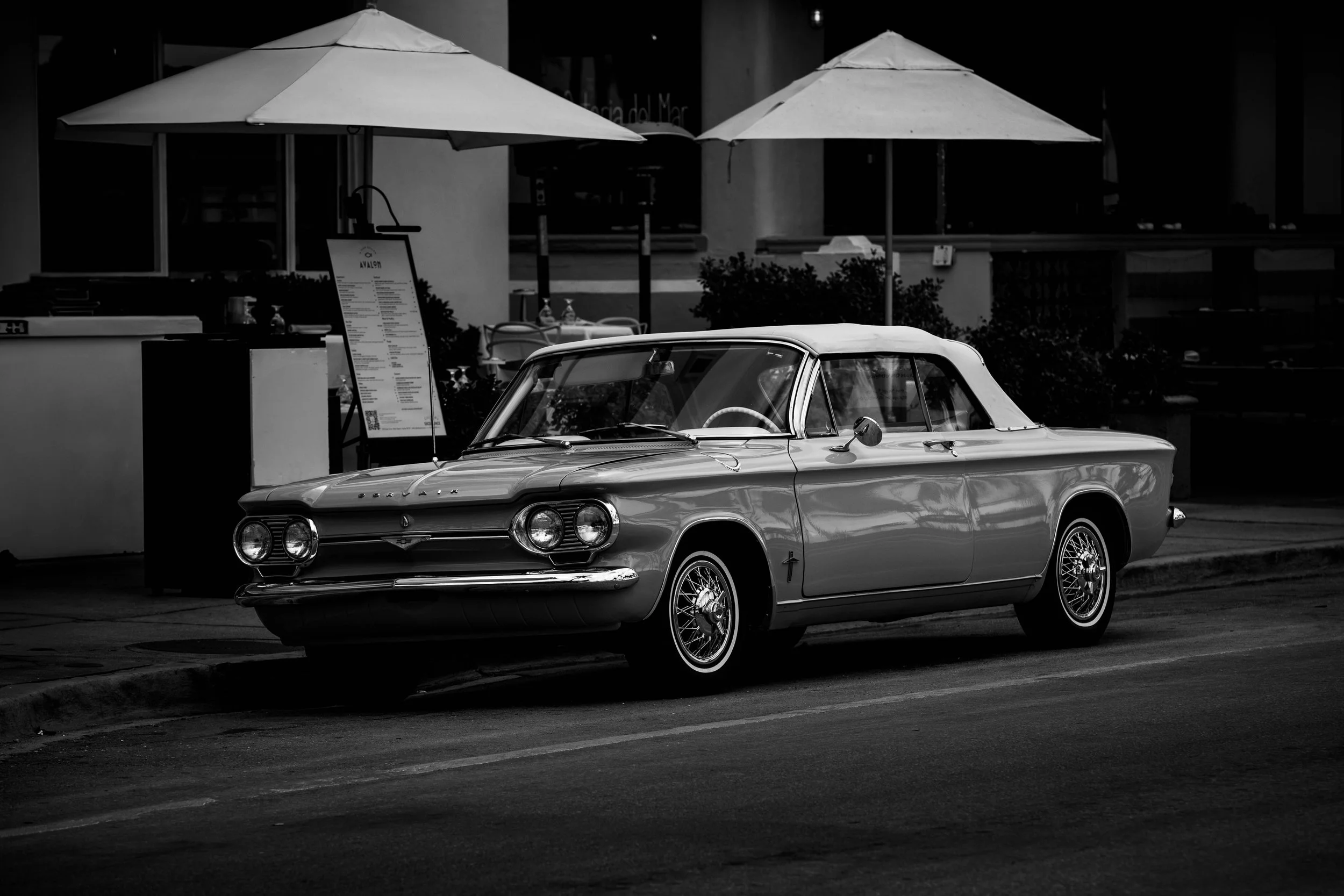 A vintage car parked on the street with two large umbrellas set up above it, casting shadows. There is a menu stand and some outdoor seating in the background, suggesting a cafe or restaurant setting.