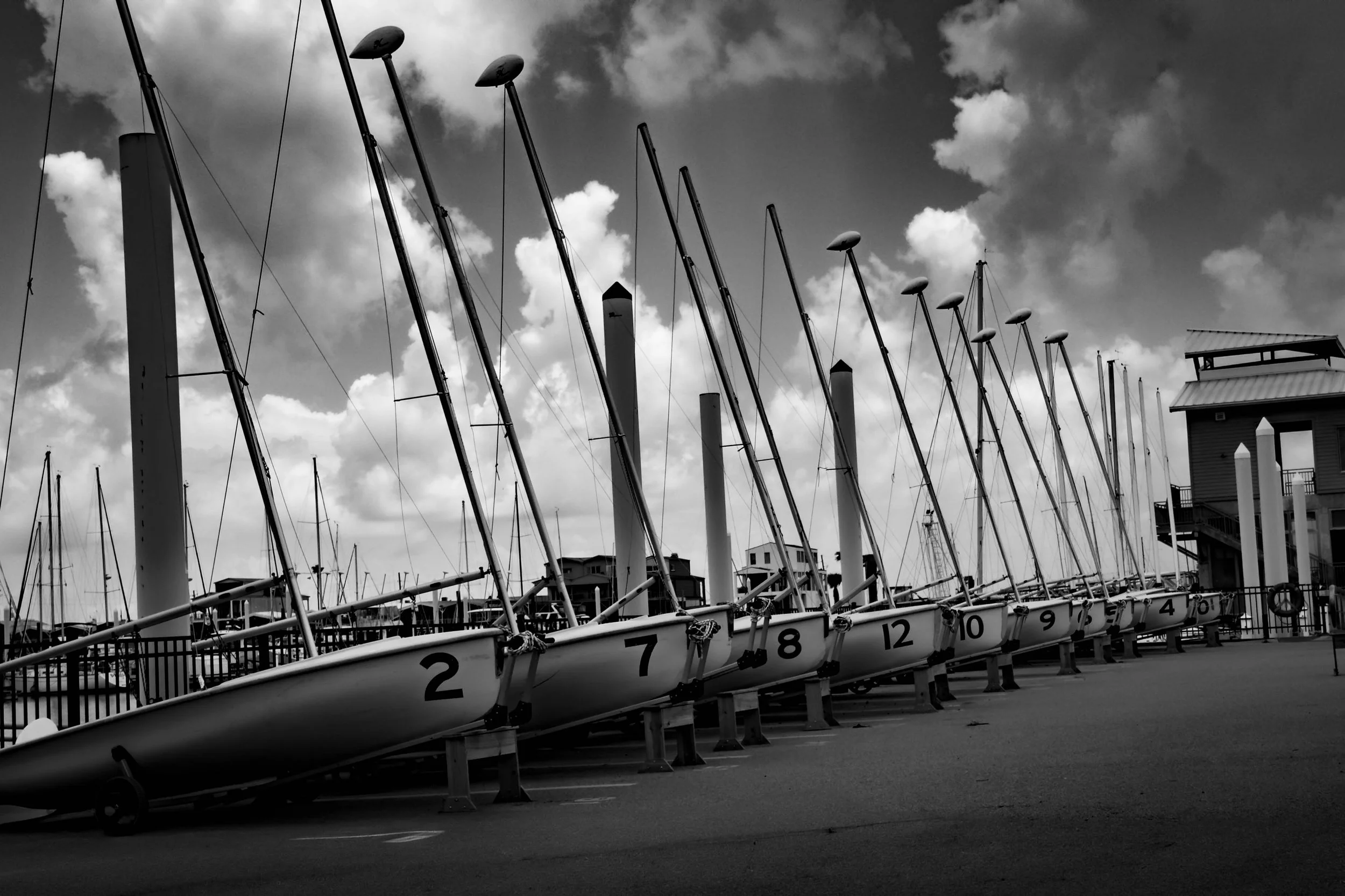 Black and white photo of sailboats with masts docked at a marina, lined up in a row with numbered hulls, under a cloudy sky.