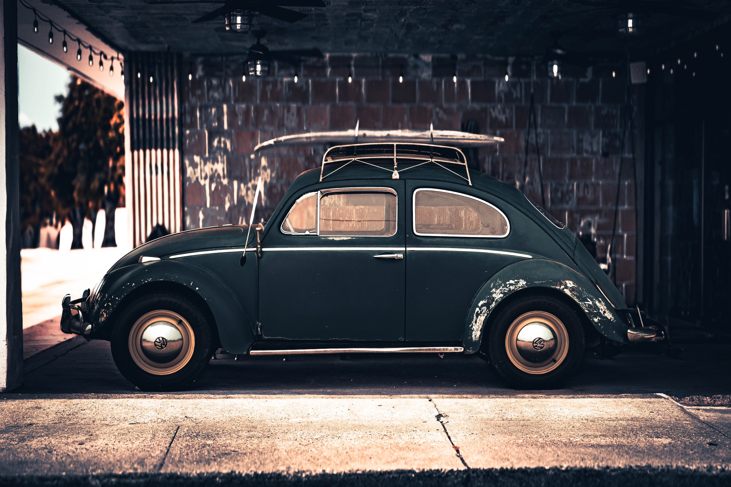 A vintage dark green Volkswagen Beetle parked in a garage with brick walls and string lights hanging from the ceiling.