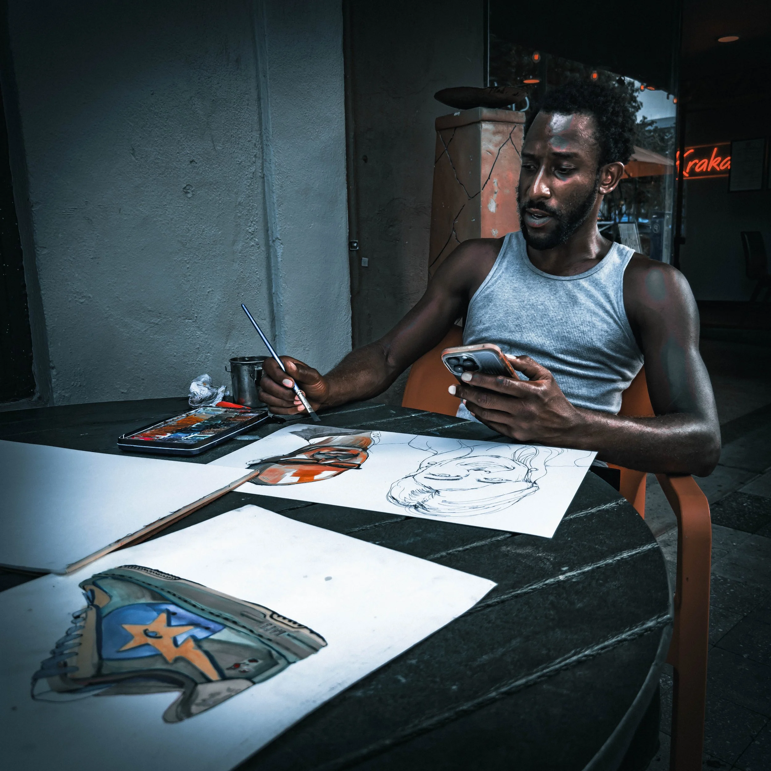 A man with curly black hair and a beard, wearing a gray sleeveless shirt, is sitting at a table in a dimly lit cafe or restaurant, looking at her phone while holding a paintbrush. There are art supplies and paper with sketches on the table, and a neo