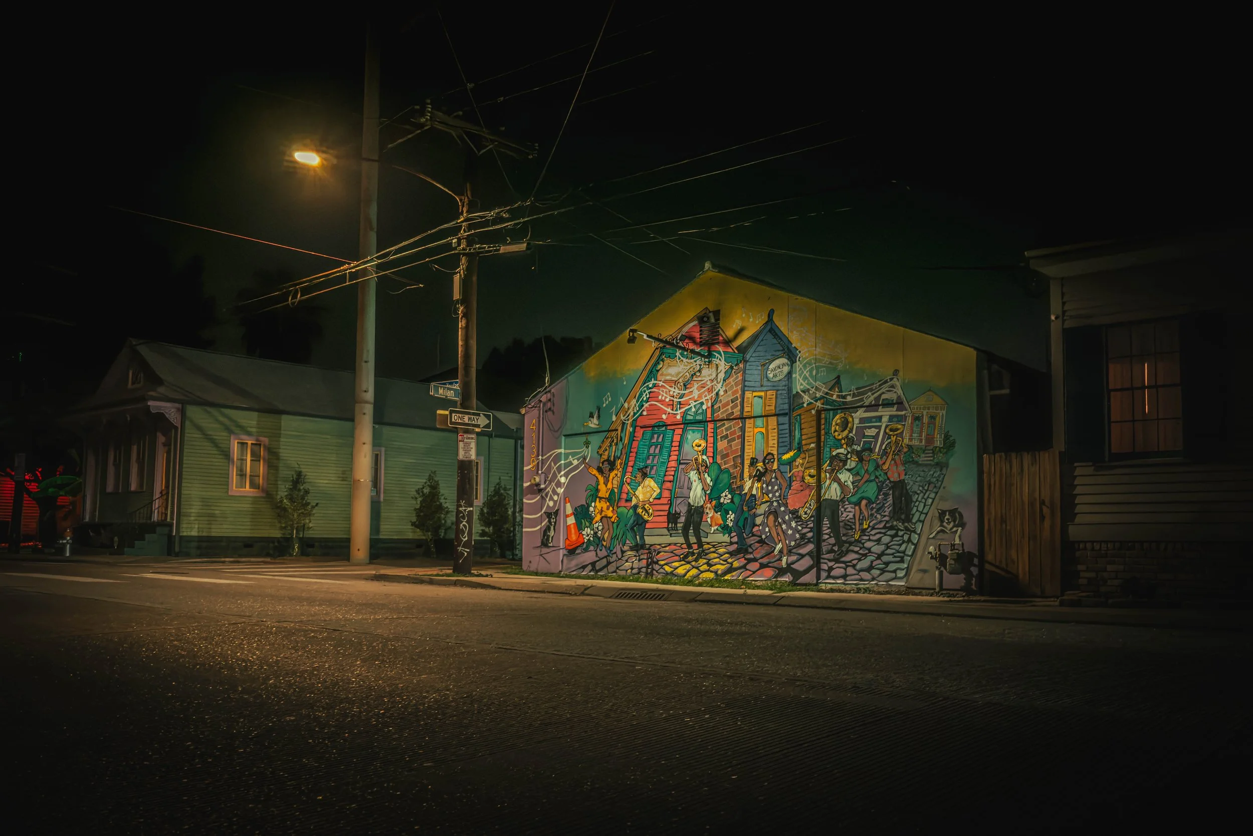 Nighttime street scene featuring a colorful mural on a building wall, with power lines and a streetlamp overhead.