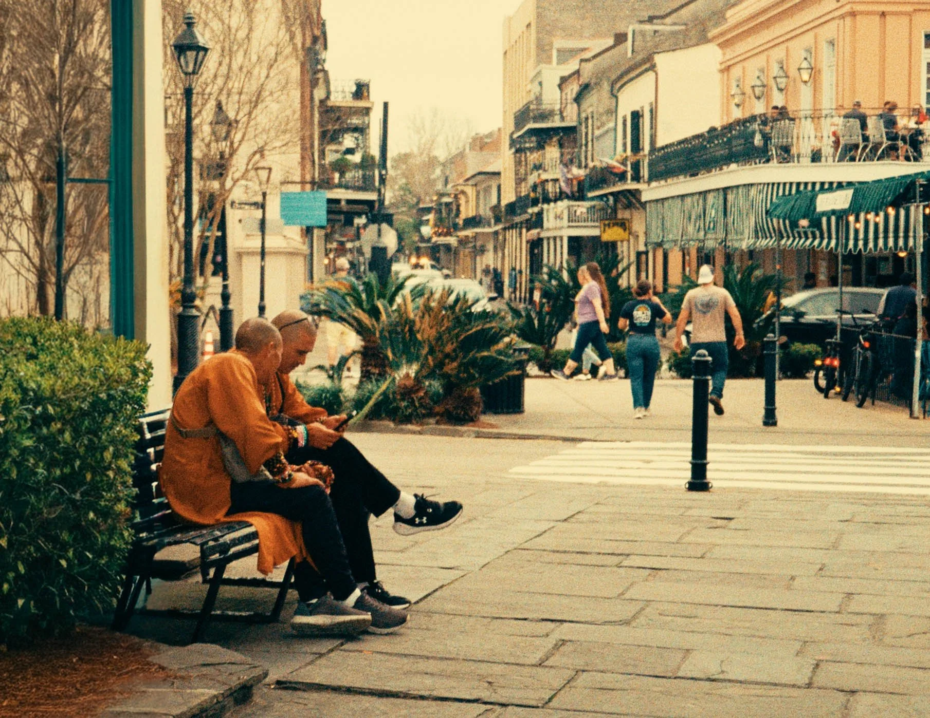 People sitting on a bench in an urban street with store fronts and pedestrians walking by.