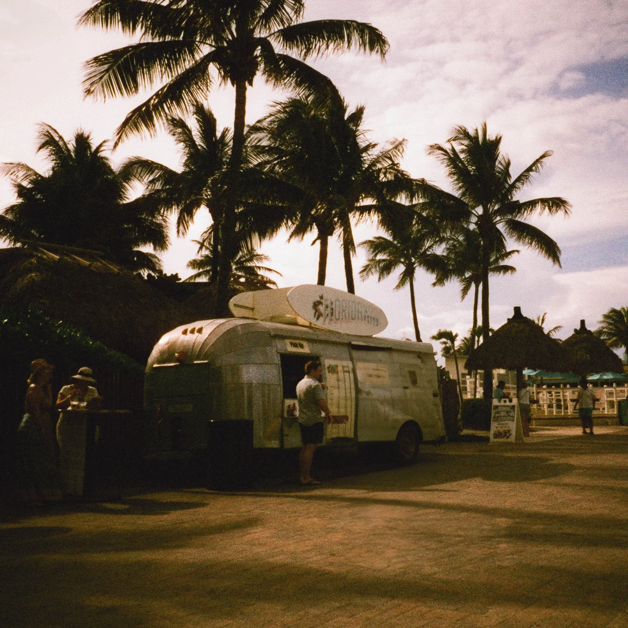 Silhouettes of palm trees against a cloudy sky during dusk with surfboards and people in the foreground.