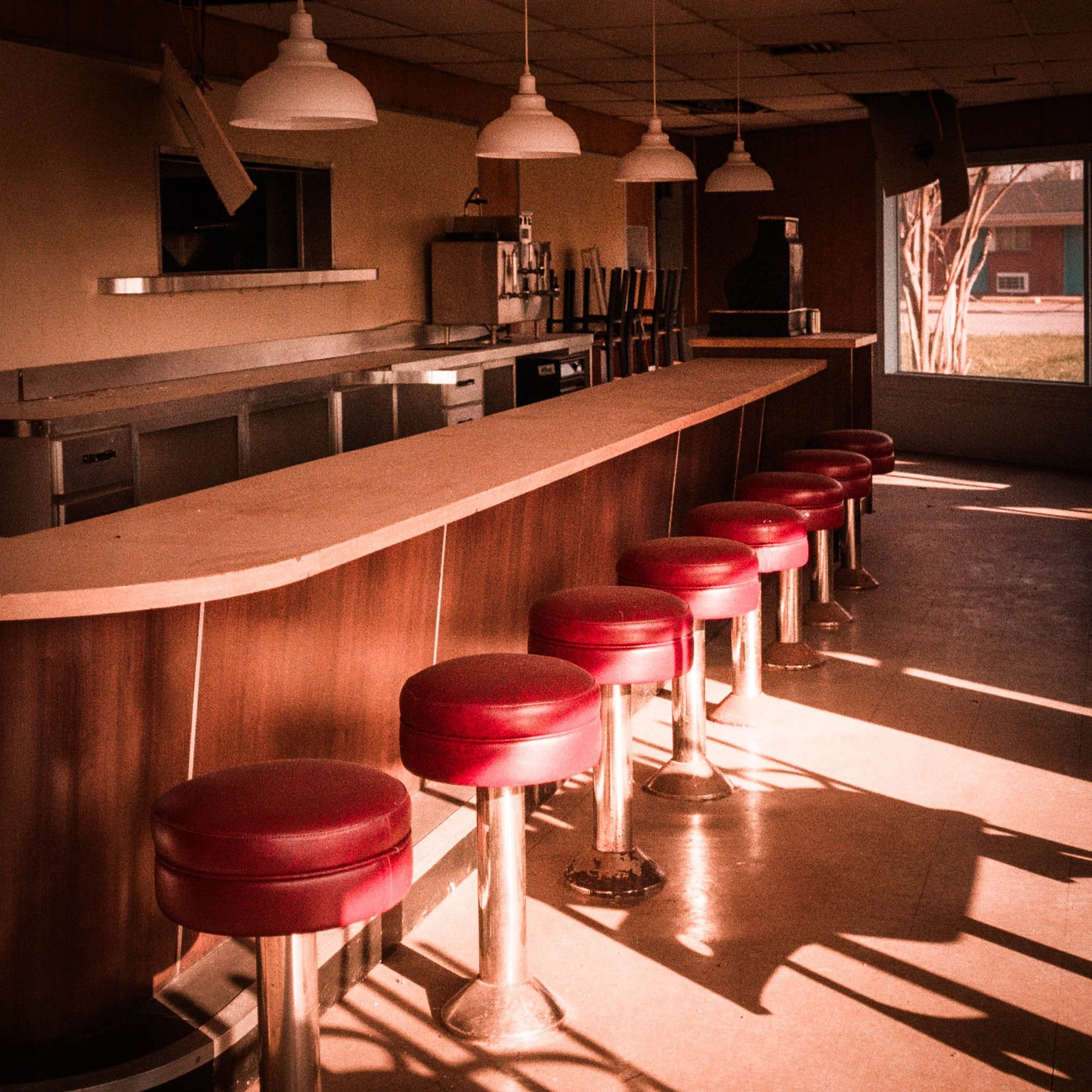 Empty restaurant or bar with a long wooden counter, red bar stools, white pendant lights, and a large window showing an outdoor scene with trees.