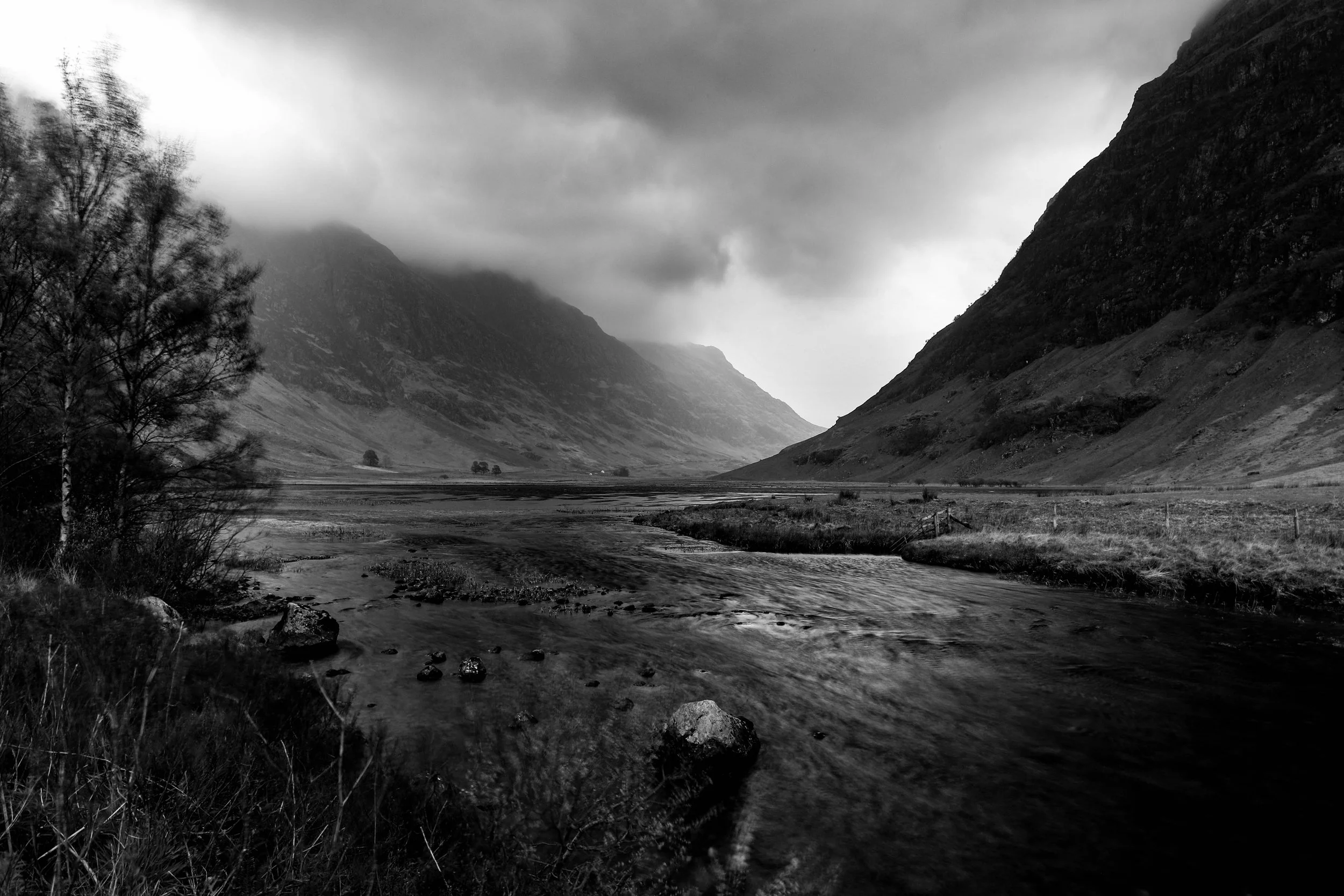 Black and white landscape photograph of a river flowing through a valley with mountains on either side, some trees in the foreground, and clouds covering the mountain peaks.
