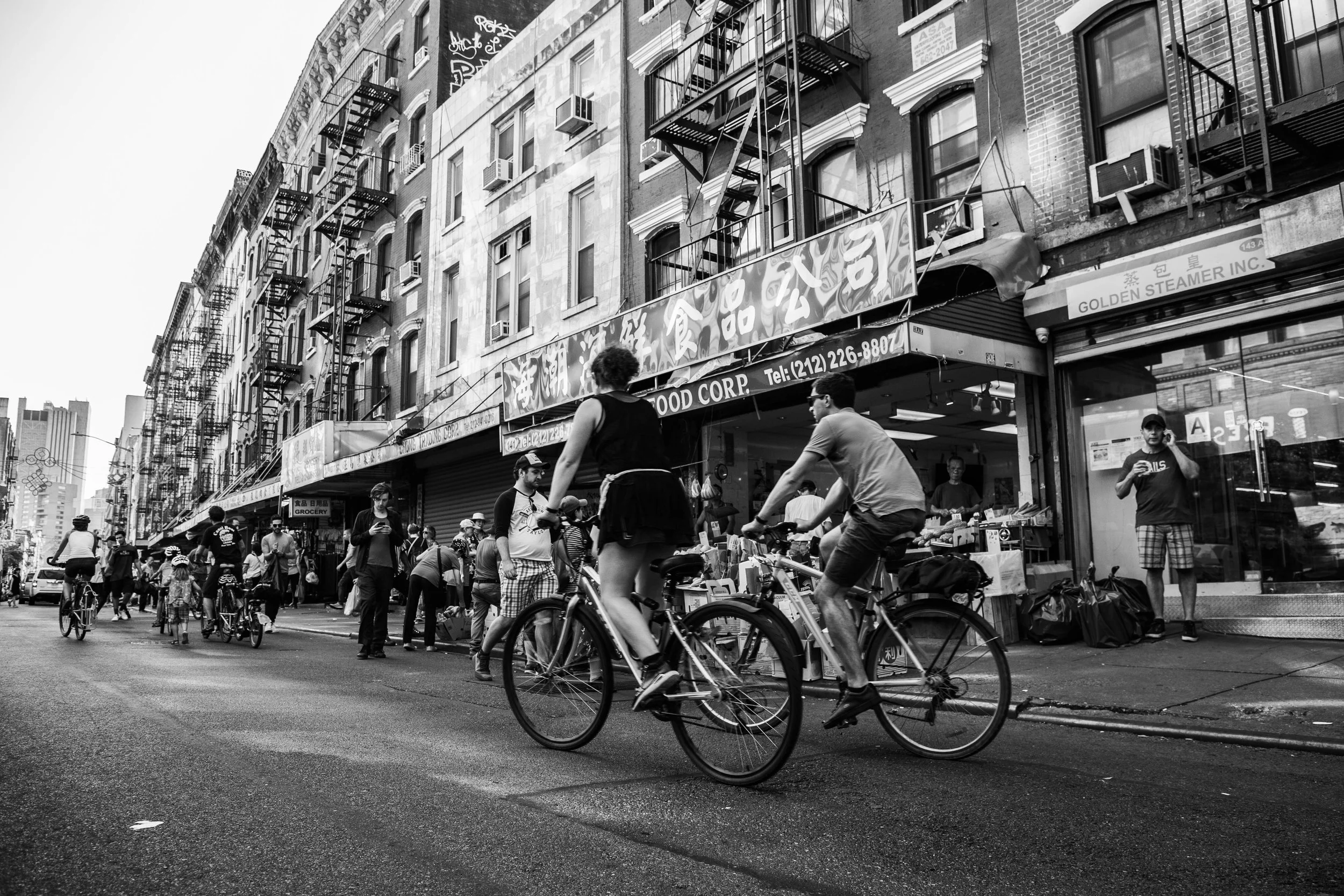 A busy city street scene with pedestrians walking and people riding bicycles. The background features multi-story brick buildings with fire escapes and air conditioning units. There are storefronts on the ground level, with one sign reading 'Grocery'