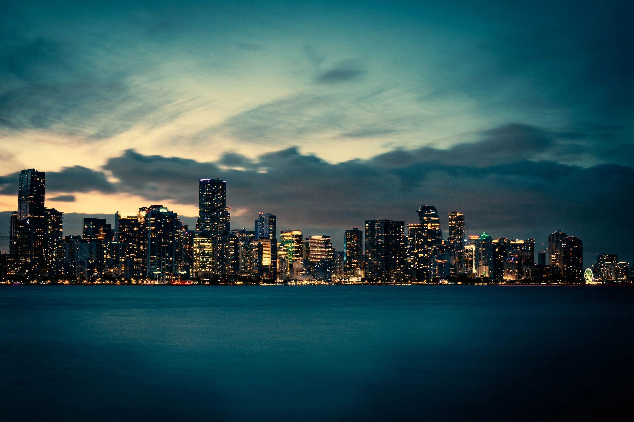 City skyline at dusk with illuminated skyscrapers and a Ferris wheel, reflected in the water in the foreground.