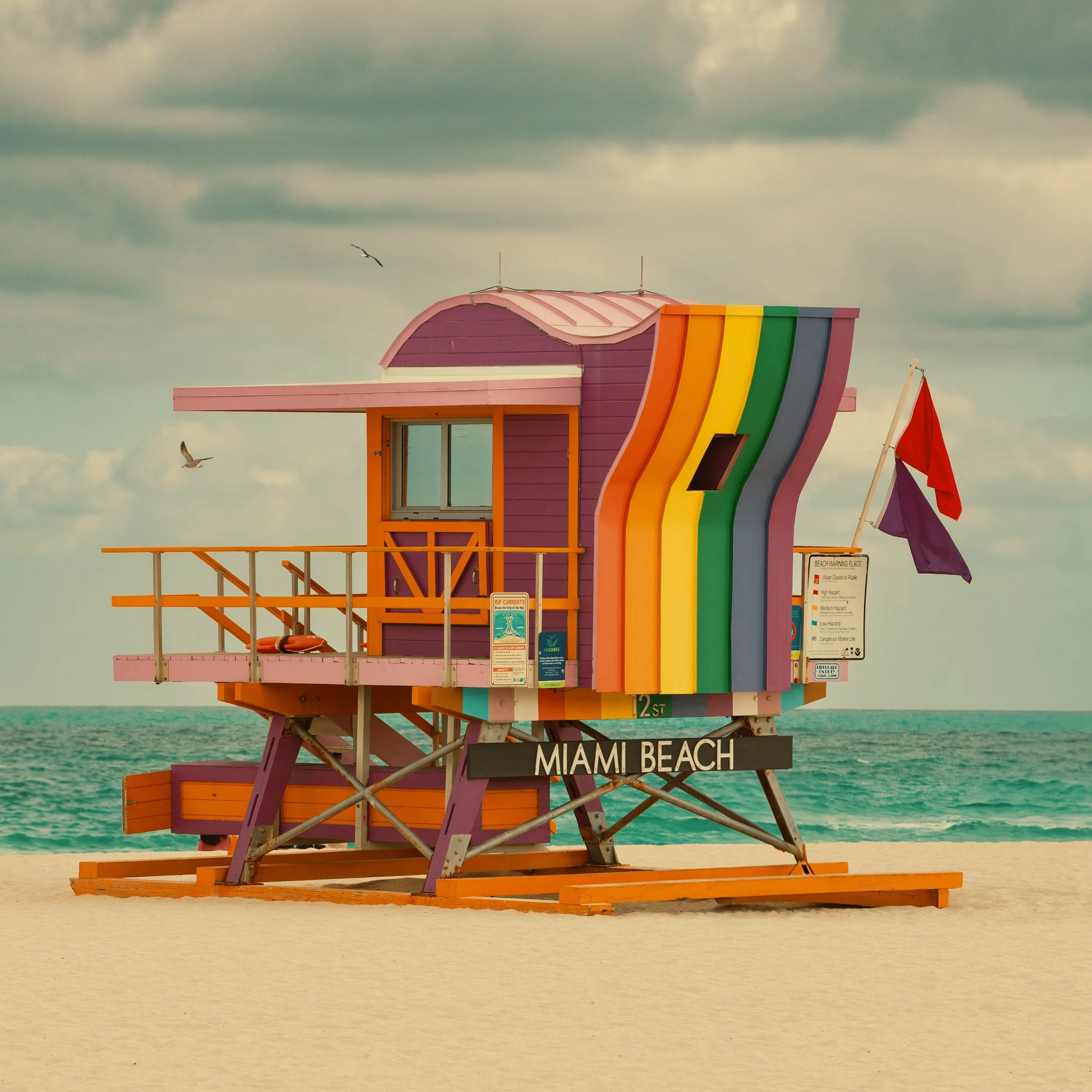 Colorful lifeguard tower with rainbow stripes on Miami Beach on a cloudy day, with seagulls flying near the tower.