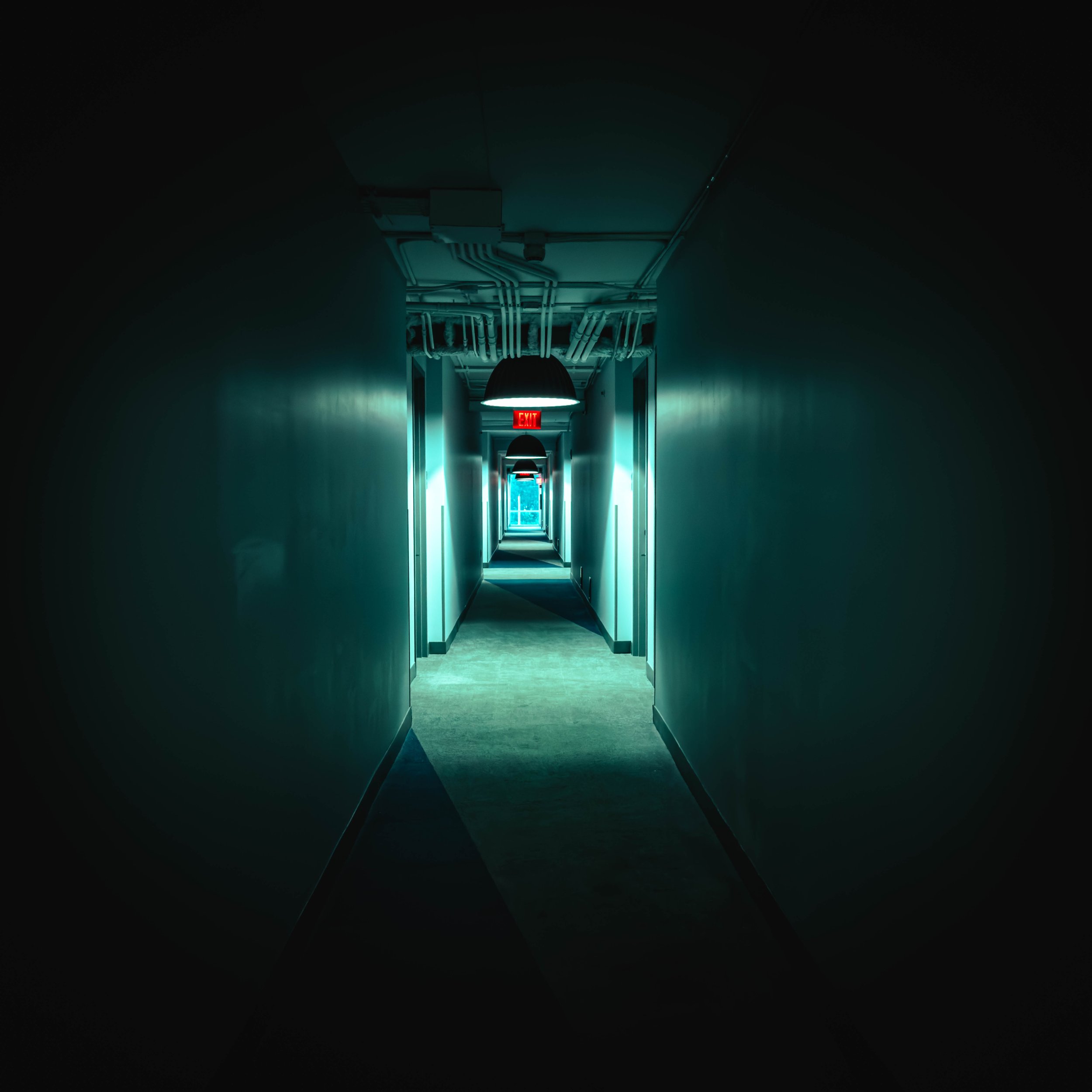 A dark hallway with teal lighting and exposed ceiling pipes, illuminated by hanging lamps and an illuminated red exit sign at the end of the corridor.