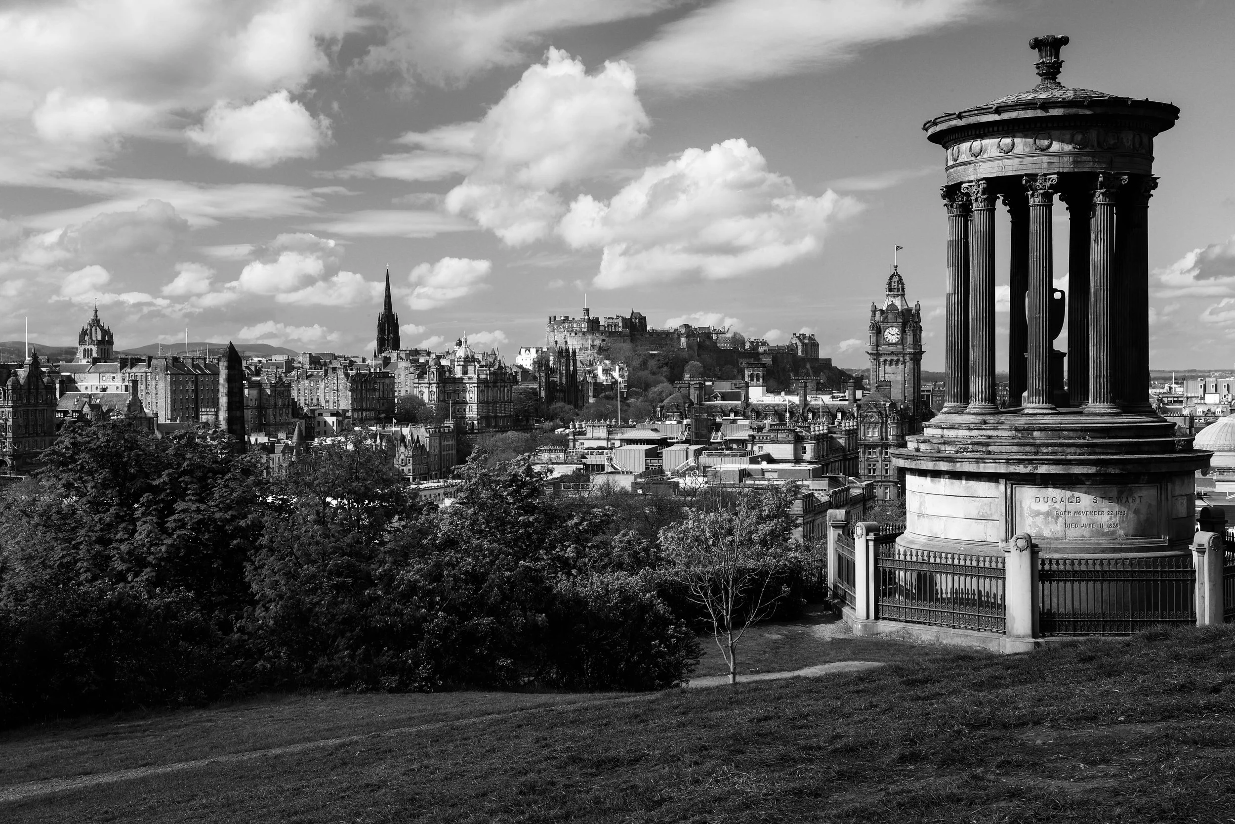 A black-and-white photo of the Edinburgh skyline, featuring the historic Edinburgh Castle on a hilltop, with a view of the city buildings and two church spires, under a cloudy sky. In the foreground, there is a monument with columns and a small fence