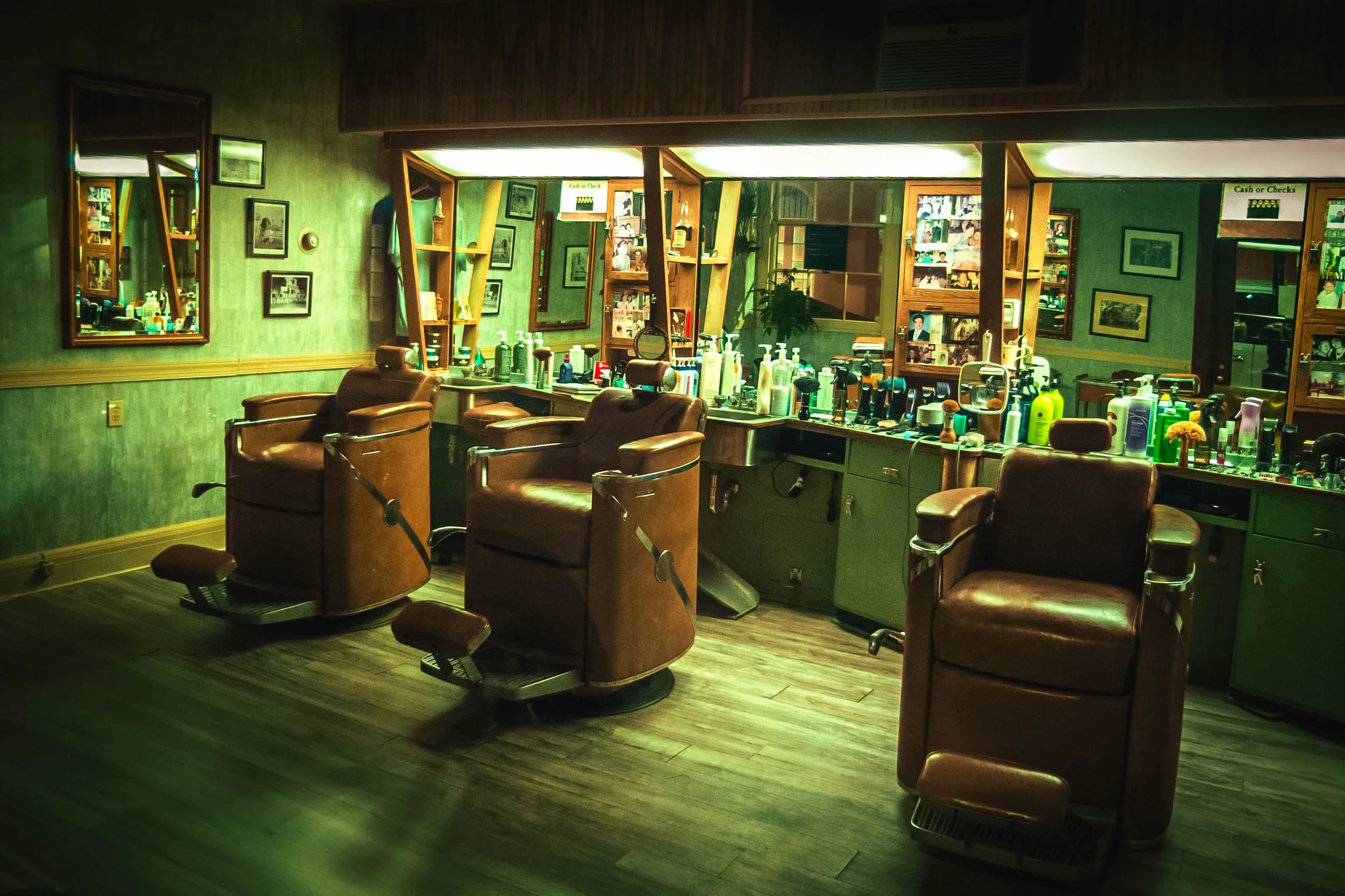 A barbershop with multiple barber chairs and mirrors along the wall, topped with various grooming products, framed pictures, and a large mirror reflecting the room.