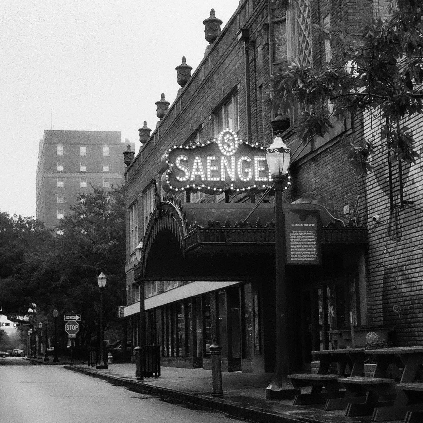A street scene featuring a building with a brick facade and a lit sign that says 'SAENGE.' The building has decorative architectural details and a small awning. There are street lamps, a stop sign, and trees along the sidewalk. The photo is black and