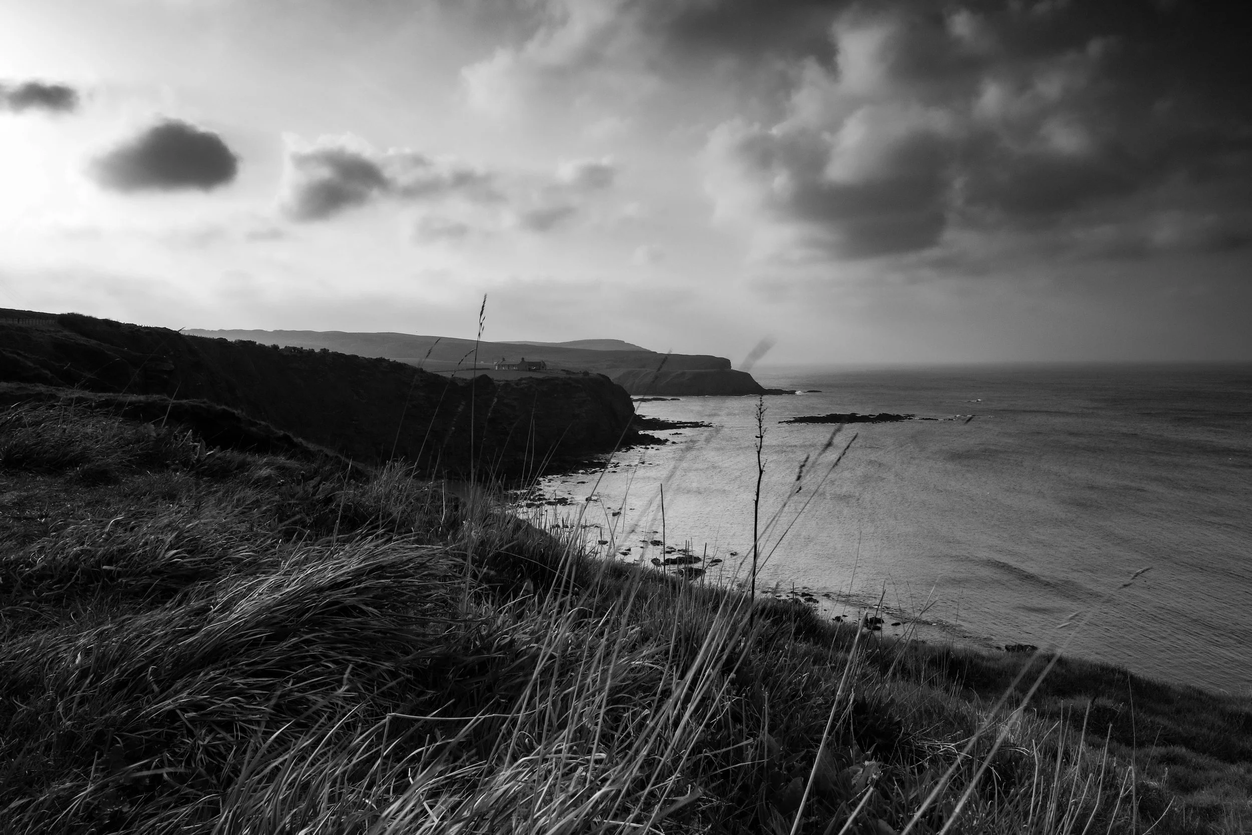 A black and white photograph of a rugged coastline with cliffs, grass in the foreground, a house or building near the cliffs in the distance, and an ocean with small waves. The sky is cloudy with dark, looming clouds.