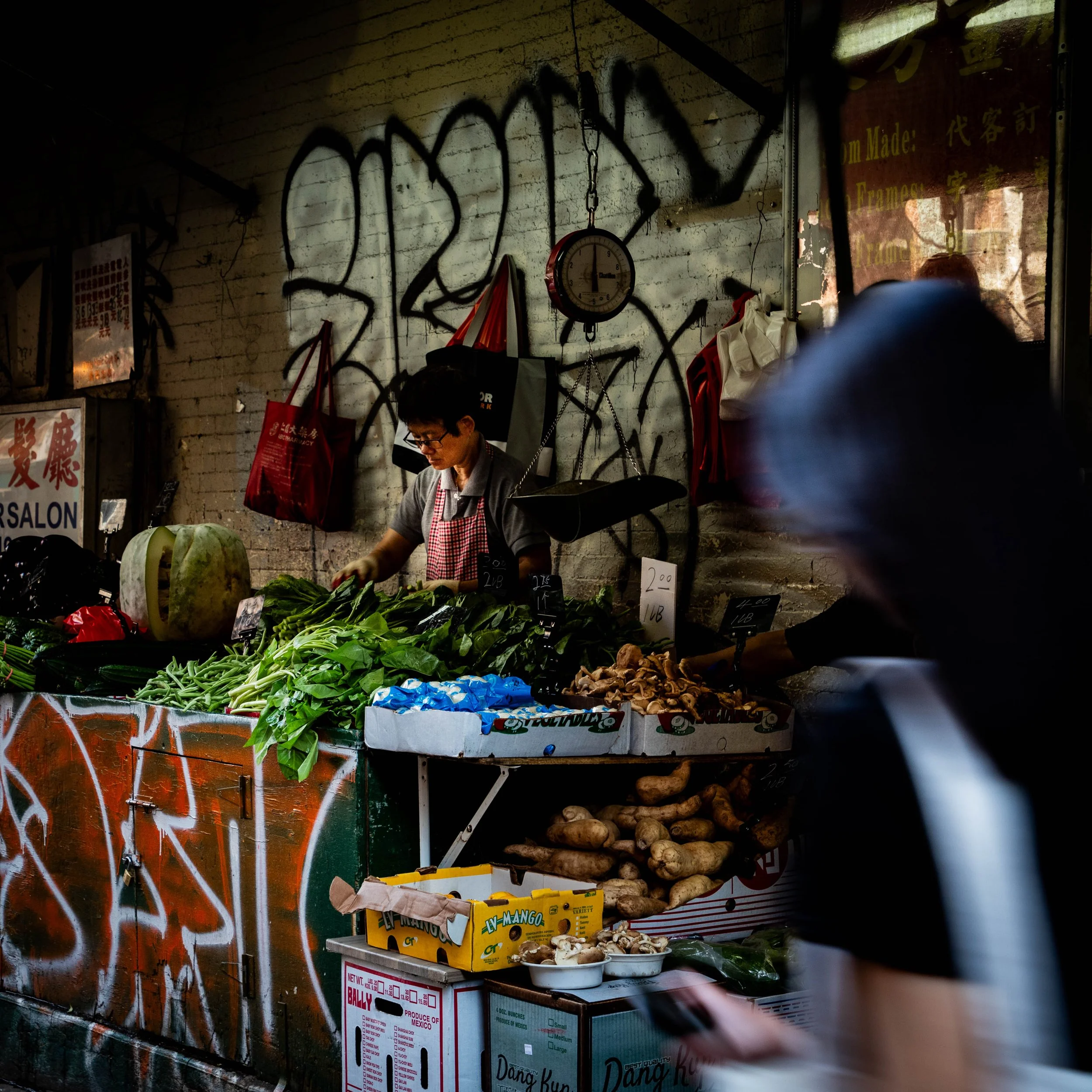 A street market stall with fresh vegetables including green beans, mushrooms, and large radish, with signs displaying prices, set against a graffiti-covered wall and a window with advertisements.