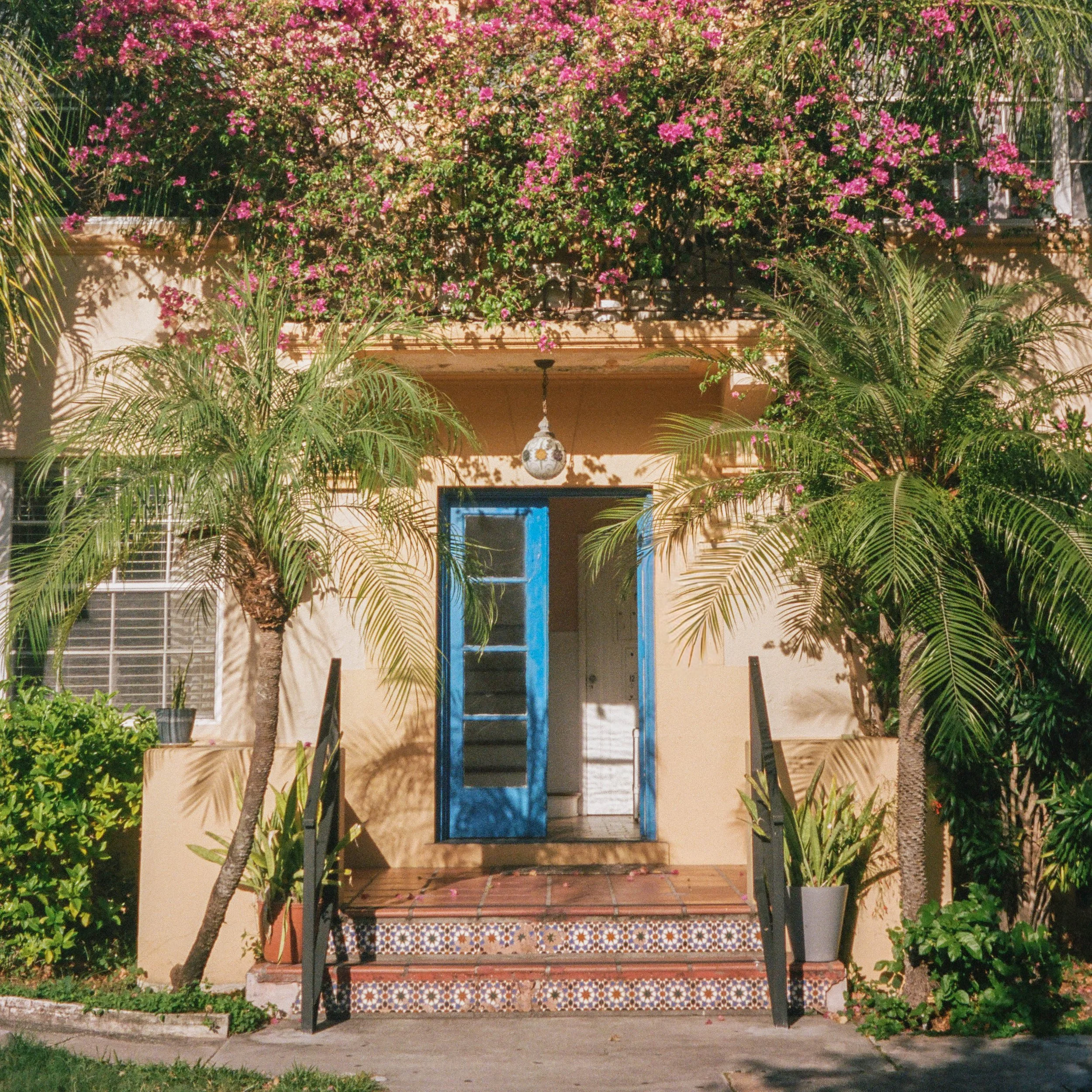 A front porch with decorative tiled steps, a hanging lantern, a blue door, lush green plants, and trees with pink flowers surrounding the area.