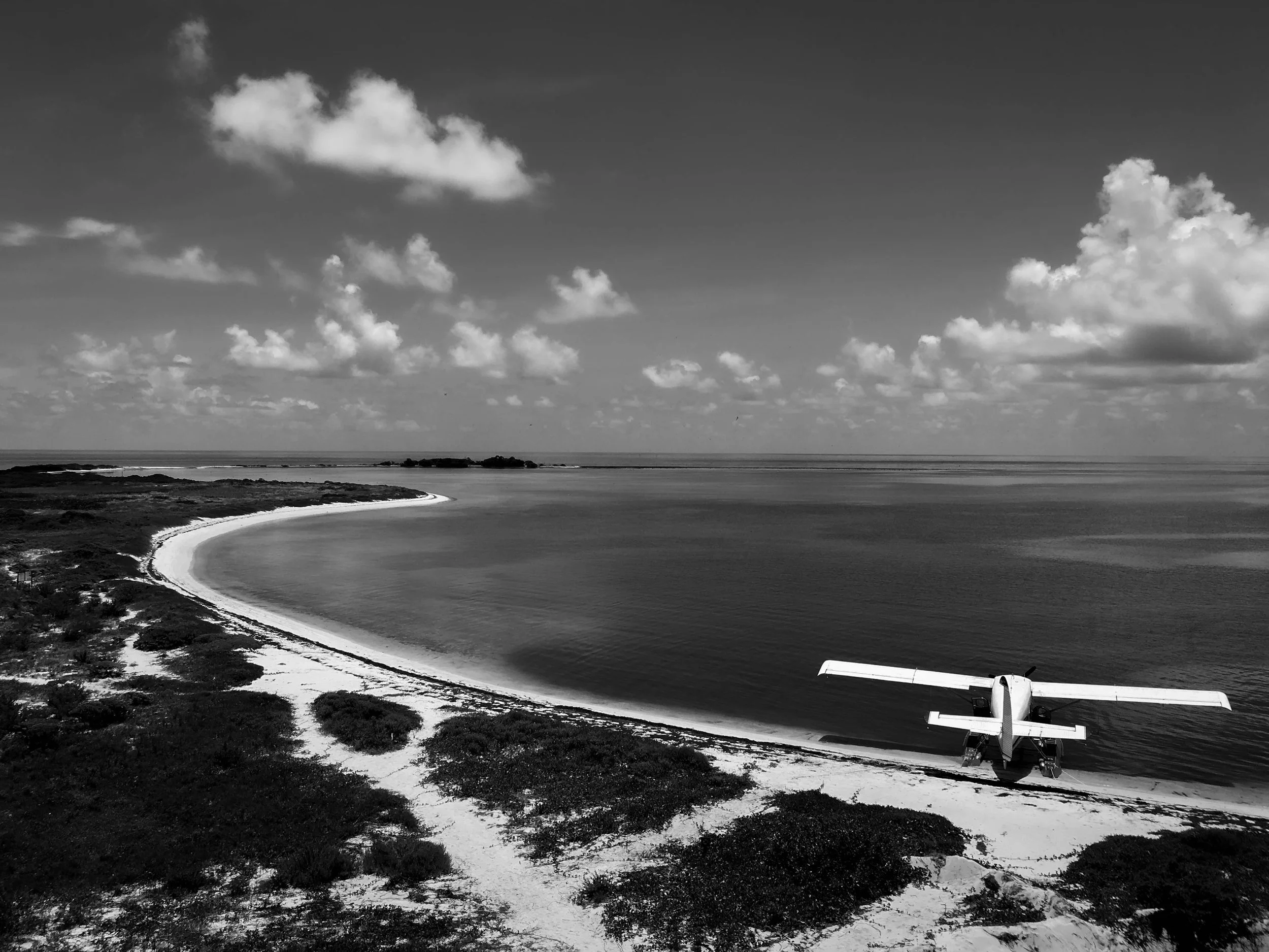 A black and white photo of a coastal landscape with a small airplane parked at the bottom right corner. There is a shoreline with sand and vegetation, calm water, and a sky with scattered clouds.