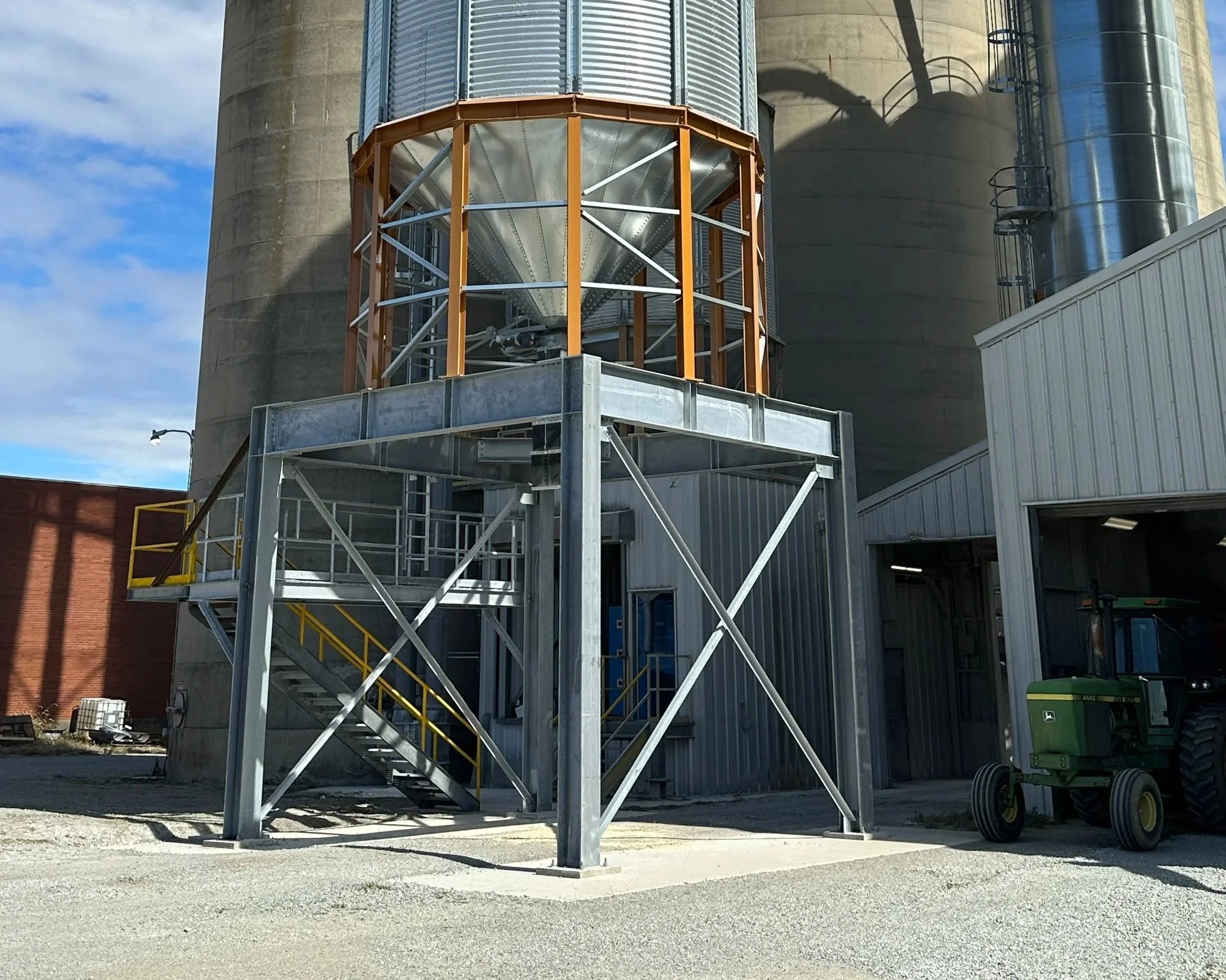 Industrial grain storage silos with metal staircase and platform, a green John Deere tractor parked nearby, and gravel ground under a partly cloudy sky.