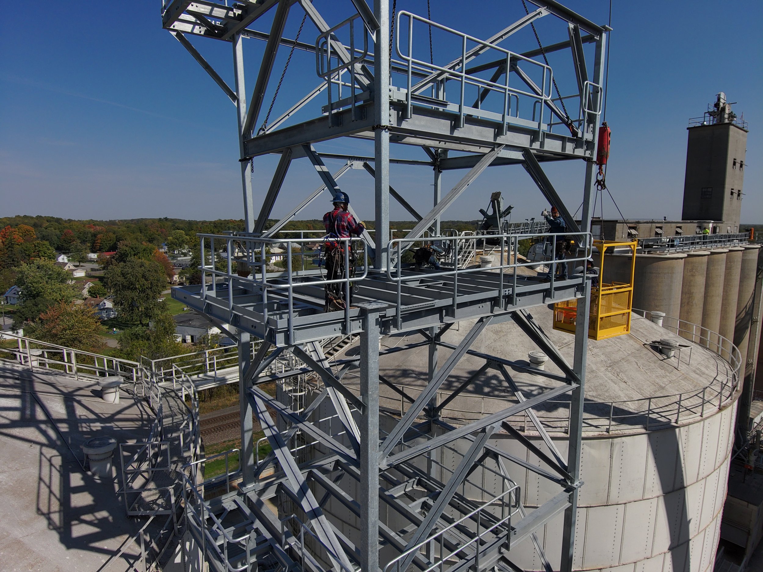 View of a large industrial structure with two workers on a metal platform. The structure includes a tall tower with a crane, railings, and ladders, set against a backdrop of trees and a clear blue sky.