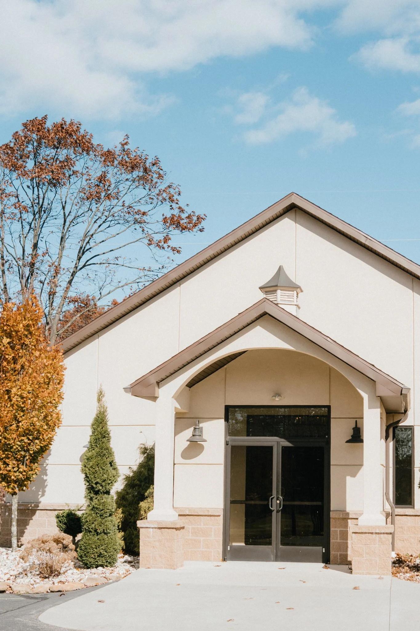 Front entrance of a beige building with a gabled roof, glass double doors, and exterior lights, surrounded by autumn trees and bushes under a blue sky.