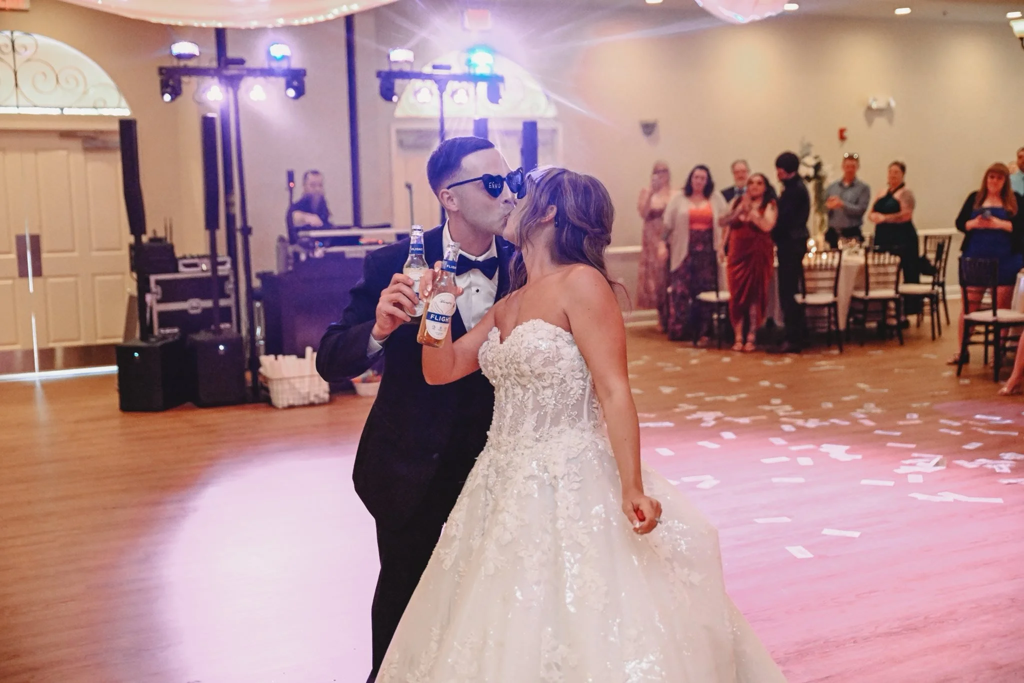 A newlywed couple sharing a kiss at their wedding reception in a decorated hall. The groom wears a black tuxedo with sunglasses, and the bride wears a white lace wedding gown. The groom holds a beer bottle, and guests are watching in the background with some standing and some seated. Confetti is scattered on the dance floor.