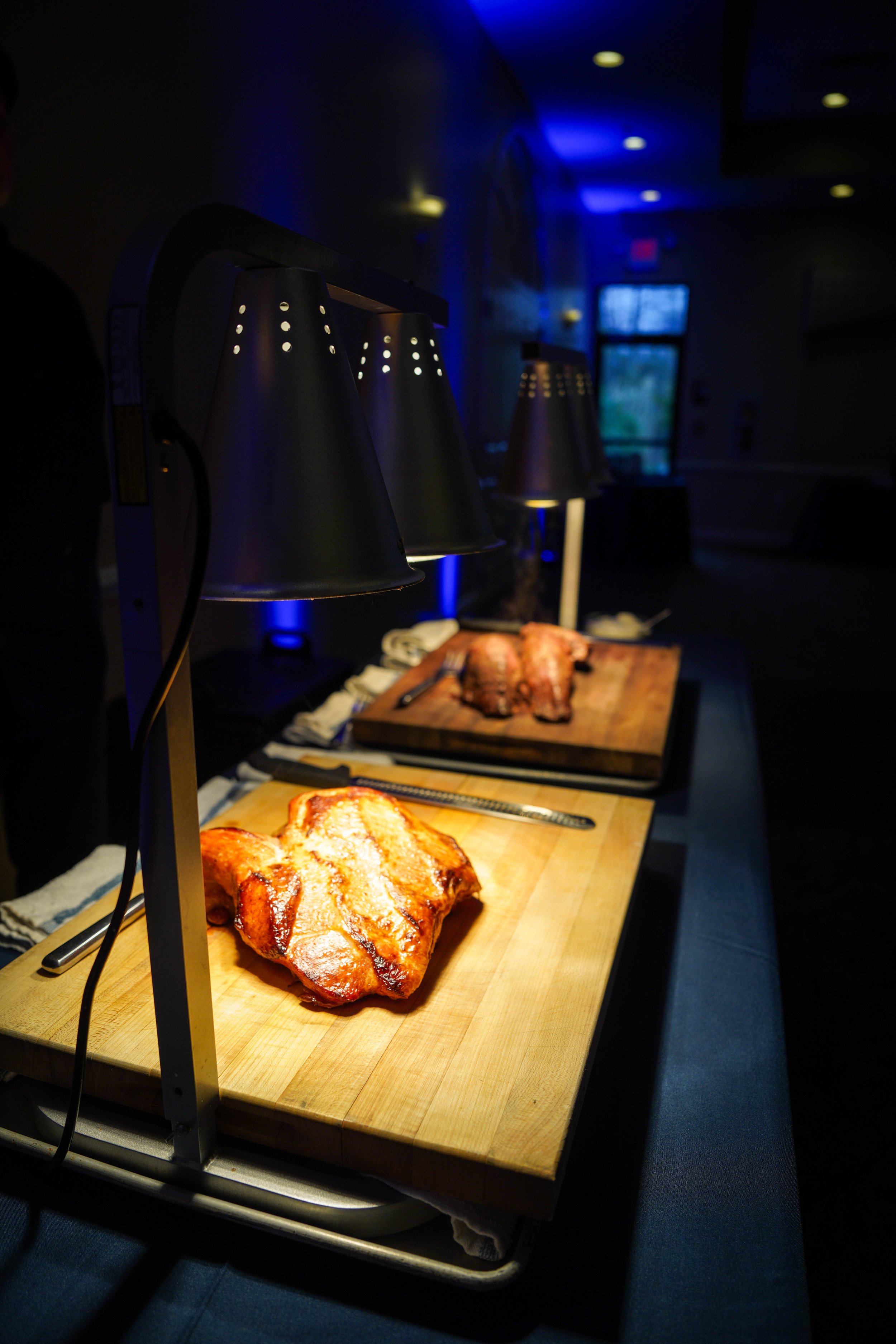 Three illuminated heat lamps above cooked meat on wooden cutting boards in a dimly lit room.