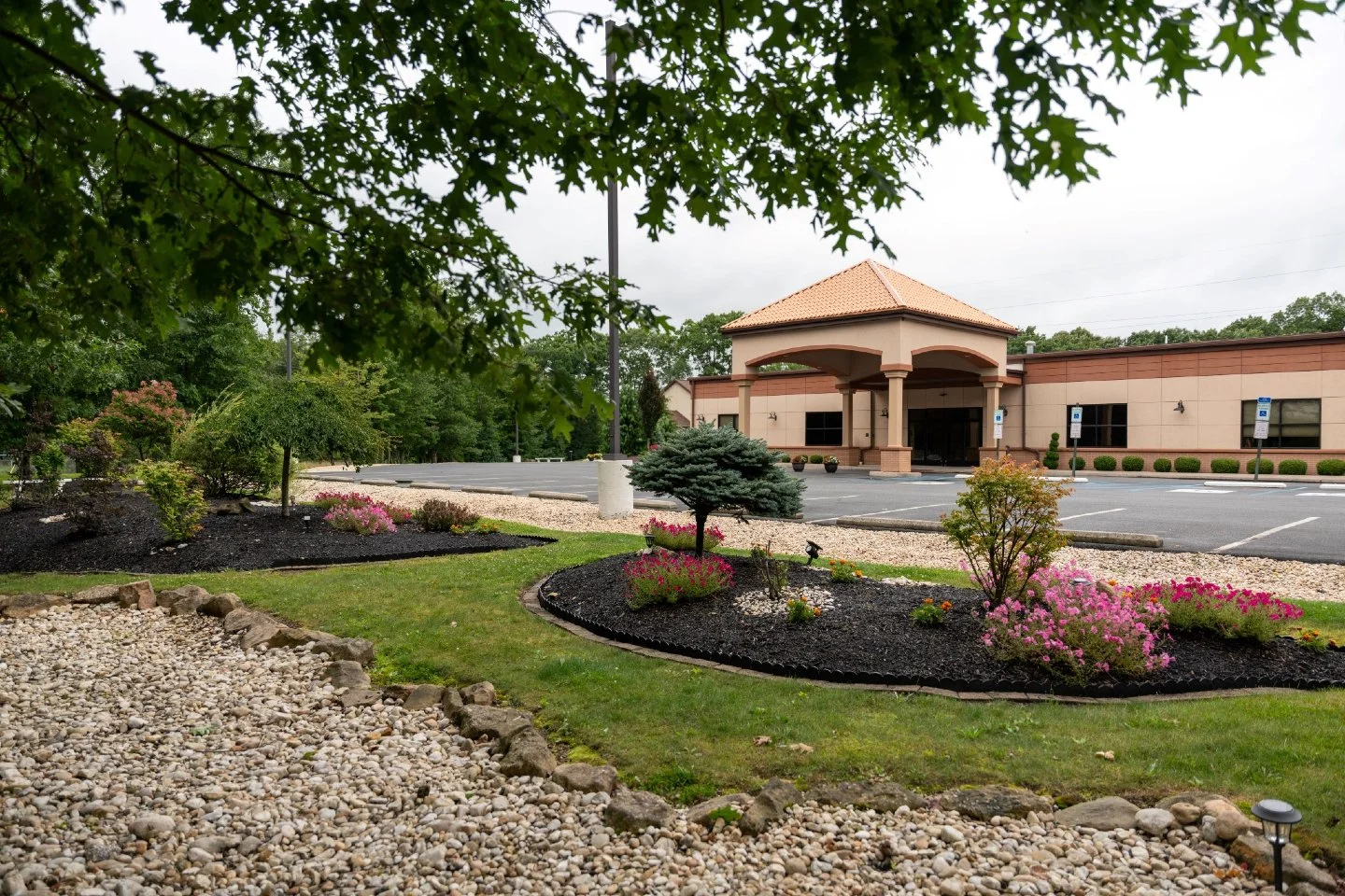 Landscaped garden with small trees, bushes, pink flowers, and rocks in front of a building with a covered entrance and a parking lot.