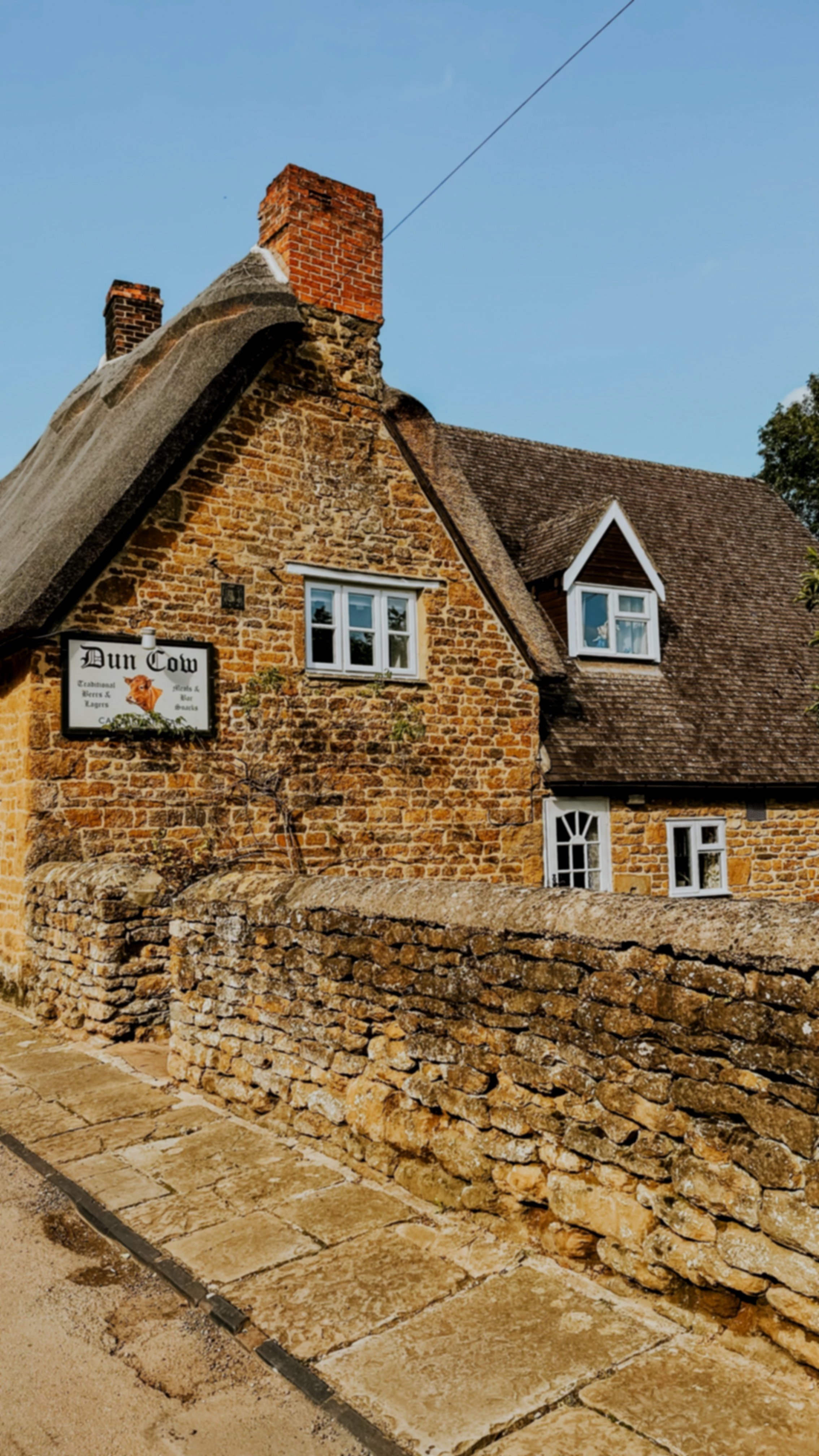 Exterior of The Dun Cow pub in Hornton, Oxfordshire, showing the cotswold stone with a traditional thatched roof, stone wall and The Dun Cow sign.