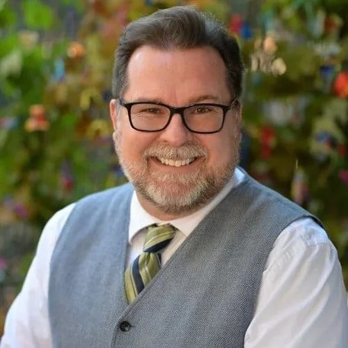 A smiling man with short grey hair, black glasses, and grey facial hair sits in front of a flower bush.