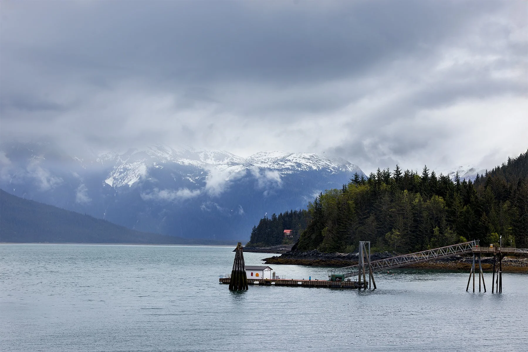 Scenic view of a dock extending into a body of water with evergreen trees and snow-capped mountains in the background under a cloudy sky.